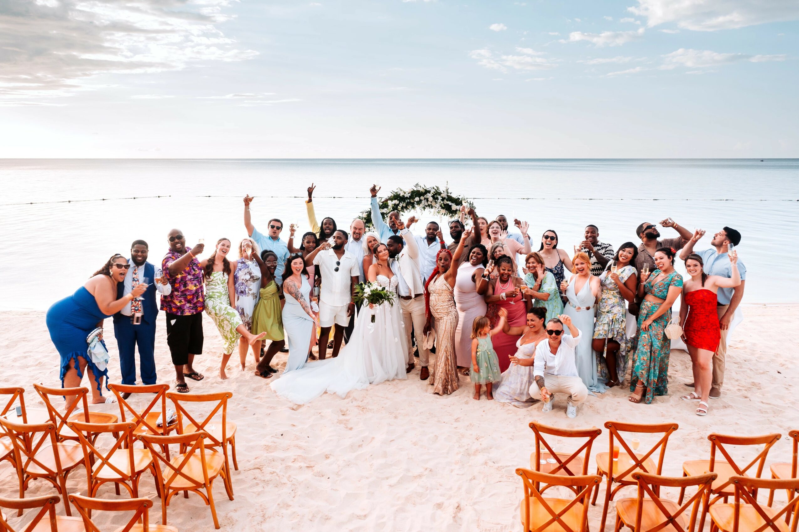 Large group of people on a beach, some in wedding attire, with chairs in the foreground and water in the background.