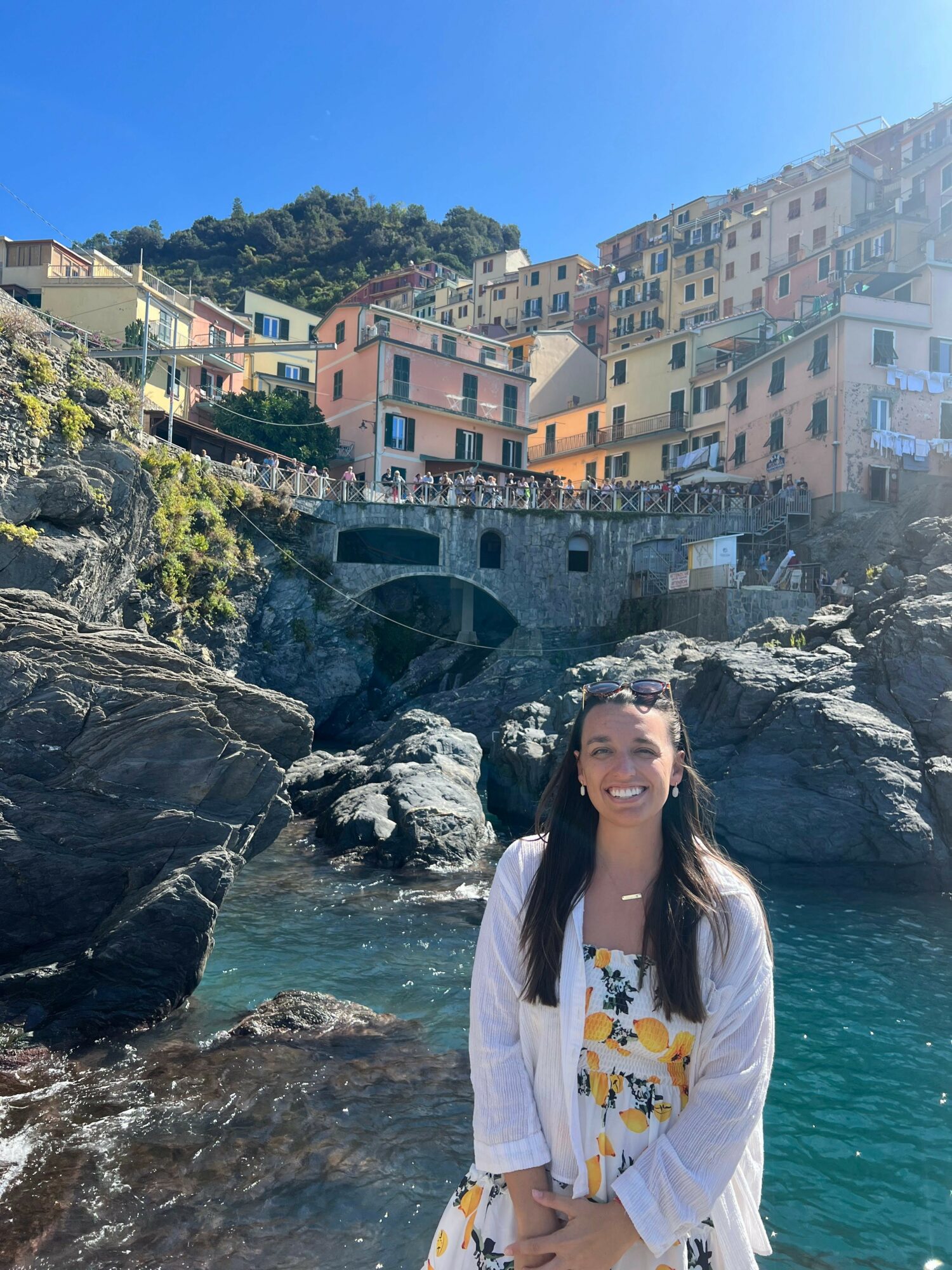 Woman standing on rocks near water with colorful hillside buildings in background.