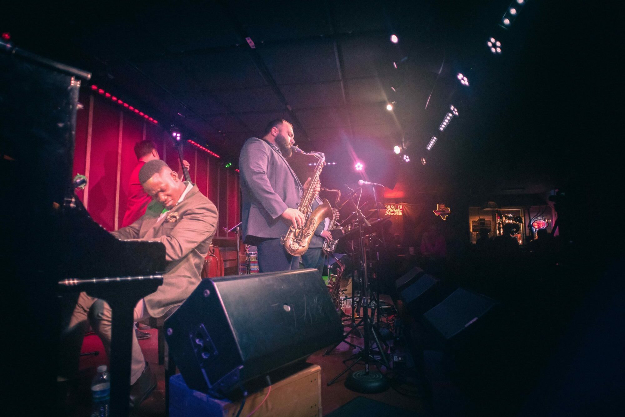 Jazz band performing on stage with a pianist, saxophonist, and microphone, illuminated by stage lights, audience in foreground.