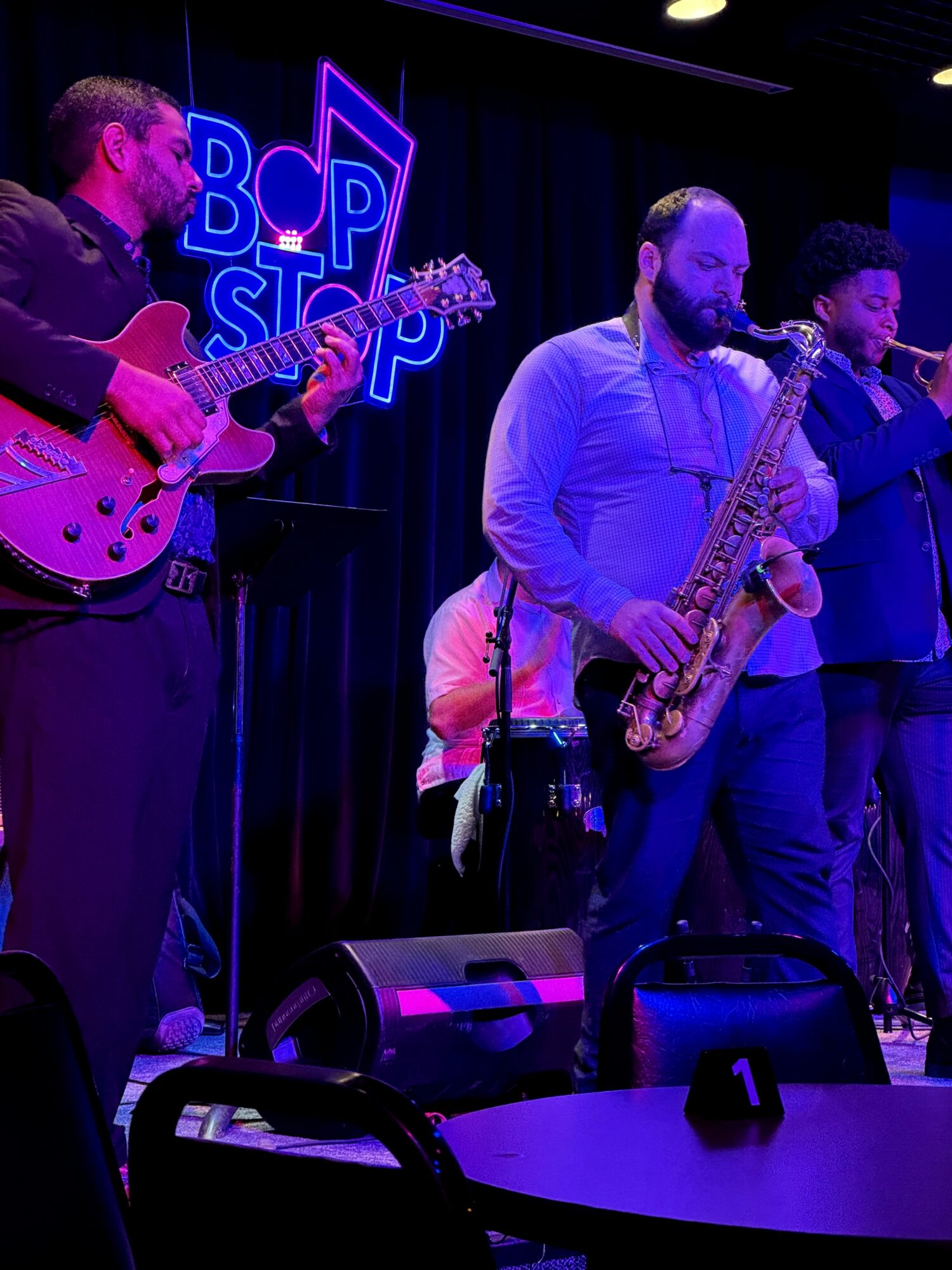 Three musicians perform on stage with a neon sign in the background, playing guitar and saxophone, in a dimly lit venue.