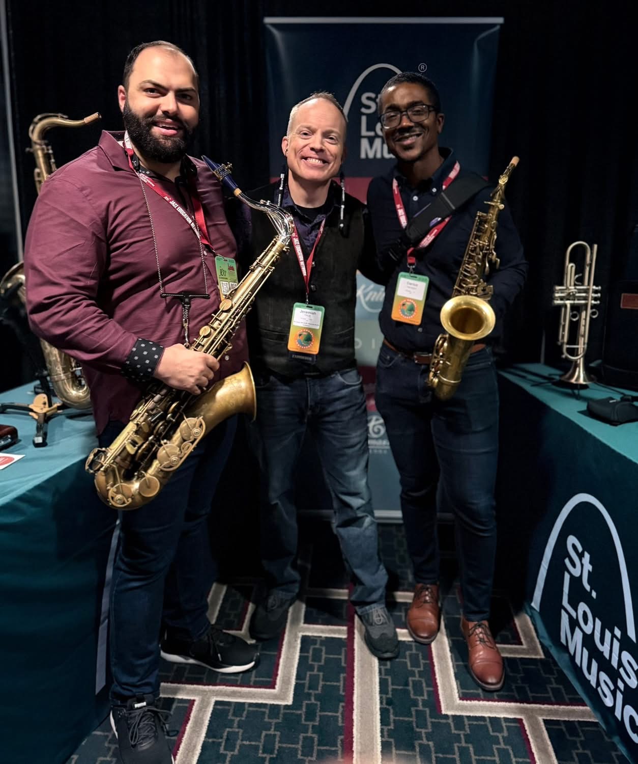 Three men smiling, holding saxophones, standing in front of a music event backdrop, with music instruments on tables nearby.