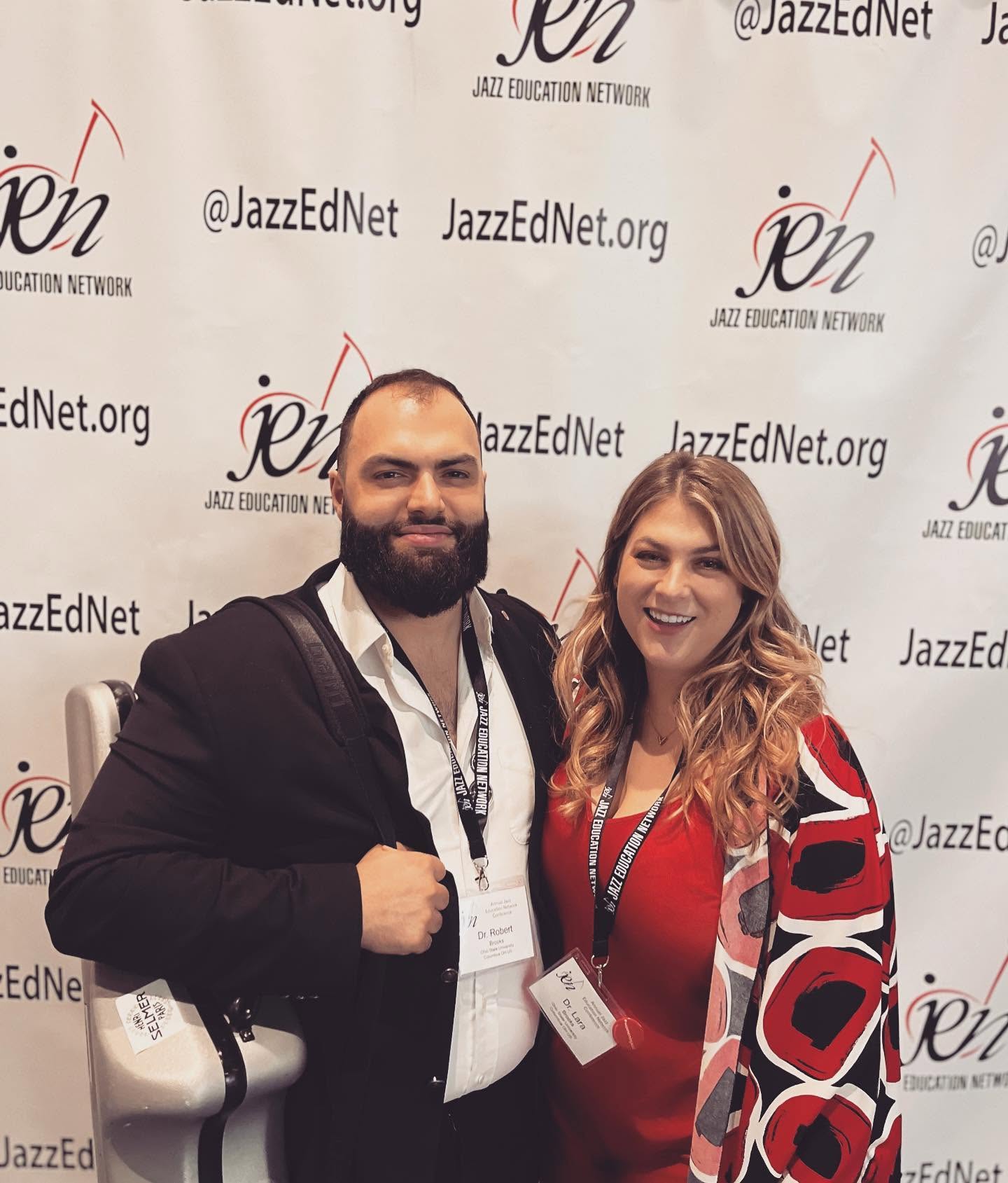 Two people smiling in front of a JazzEdNet backdrop, one with a beard and the other with long hair, wearing a red and black patterned jacket.