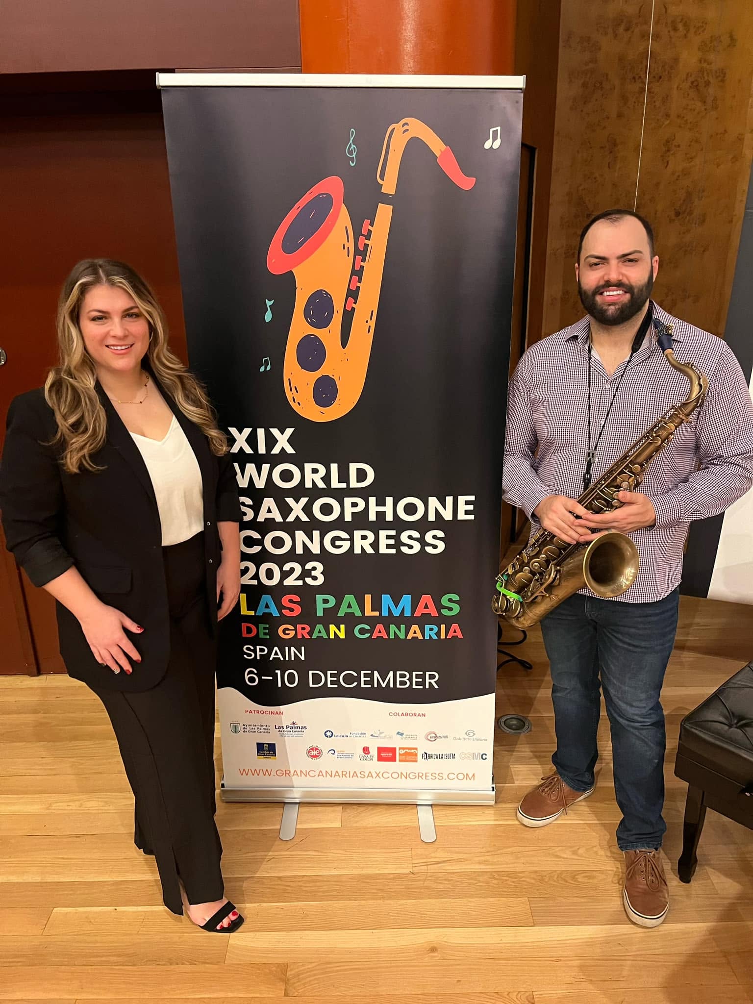 Two people standing next to a saxophone illustration on a banner at a conference, one woman and one man holding a saxophone, indoors.