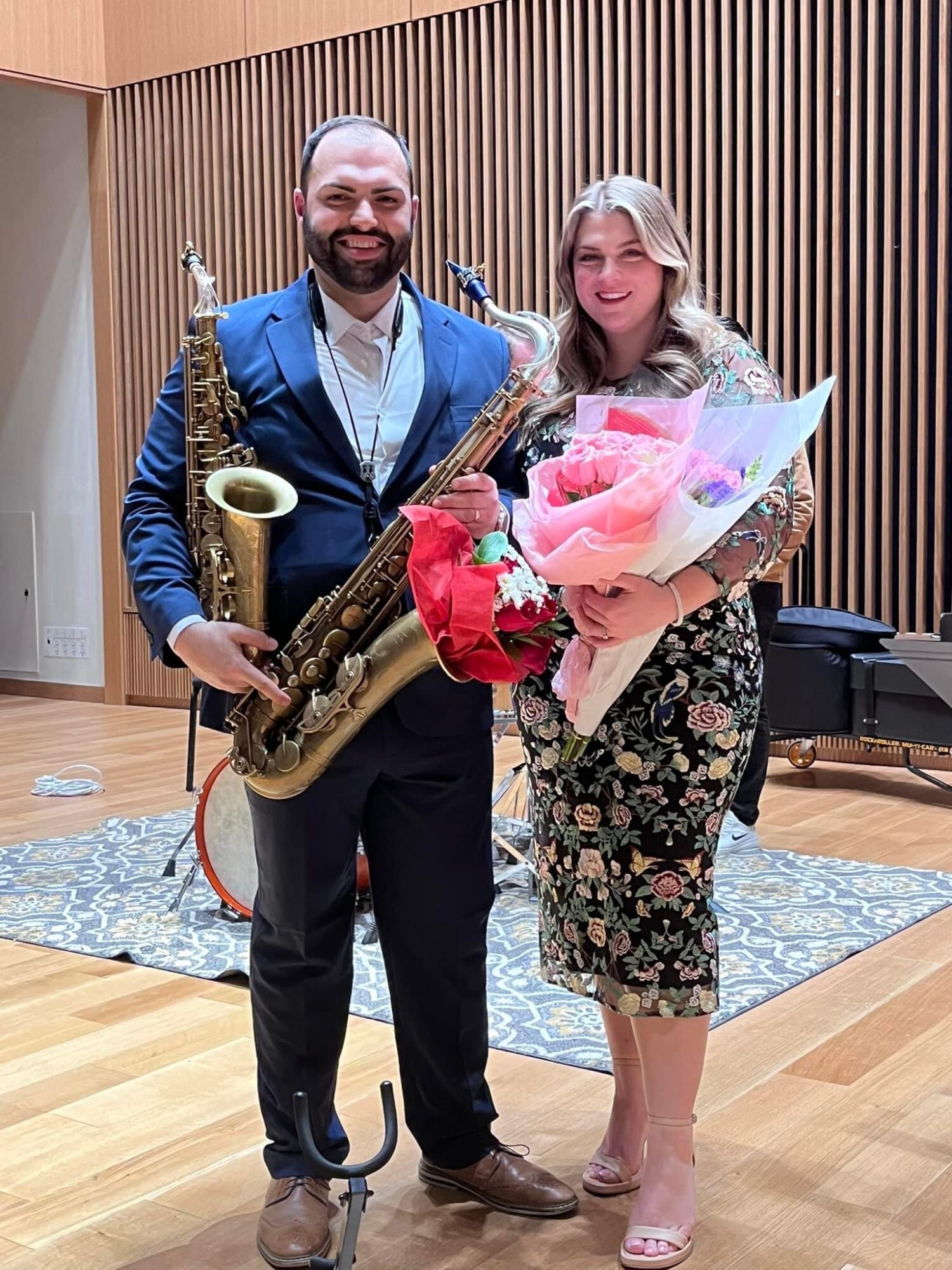 A man holding a saxophone and a woman holding a bouquet of flowers stand together in a room with wooden paneling, smiling.