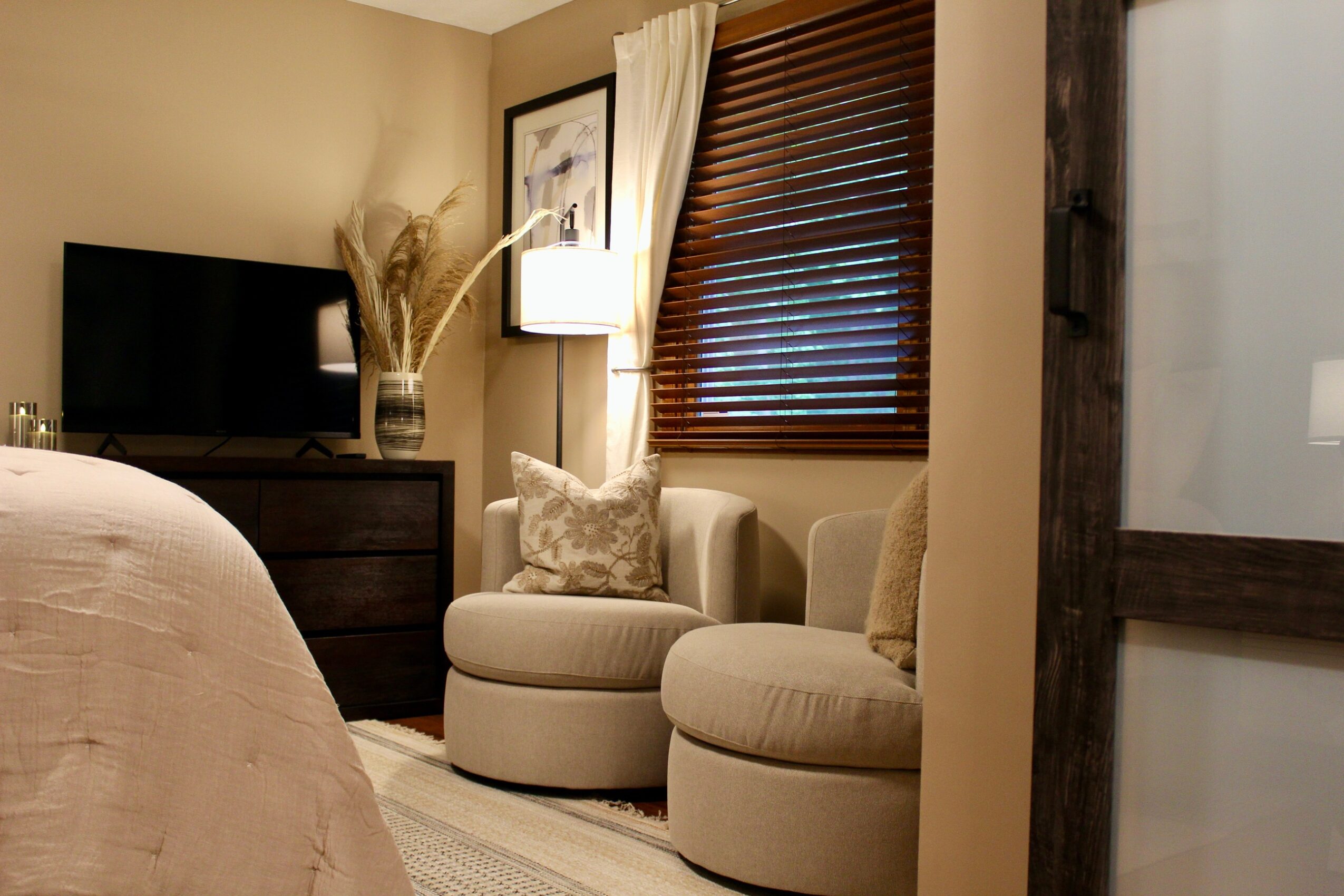 Corner of a bedroom with a TV, armchair, window with blinds, and curtains, in warm lighting.