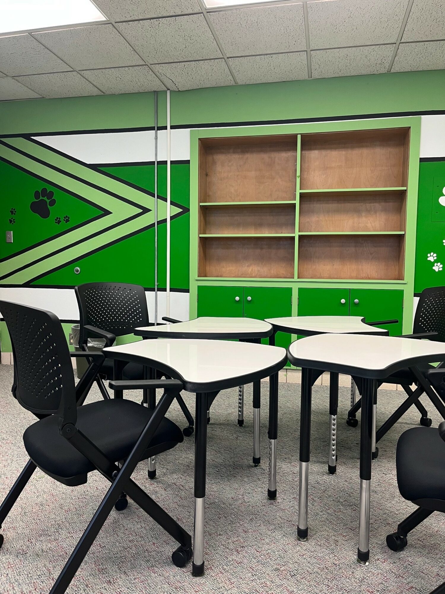 Classroom with green and black wall design, empty wooden shelves, and black chairs around white tables.