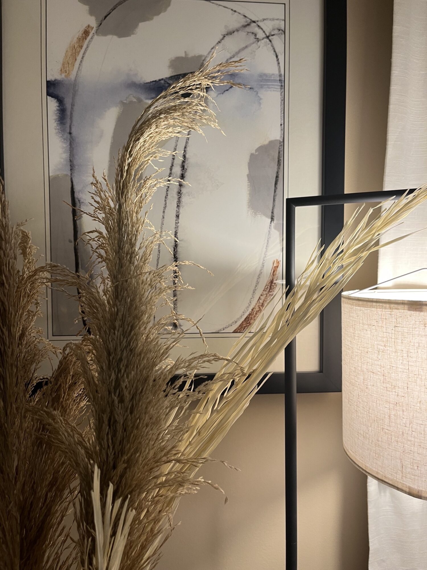 Decorative dried grasses in front of a framed abstract painting, with a lamp and curtain nearby.