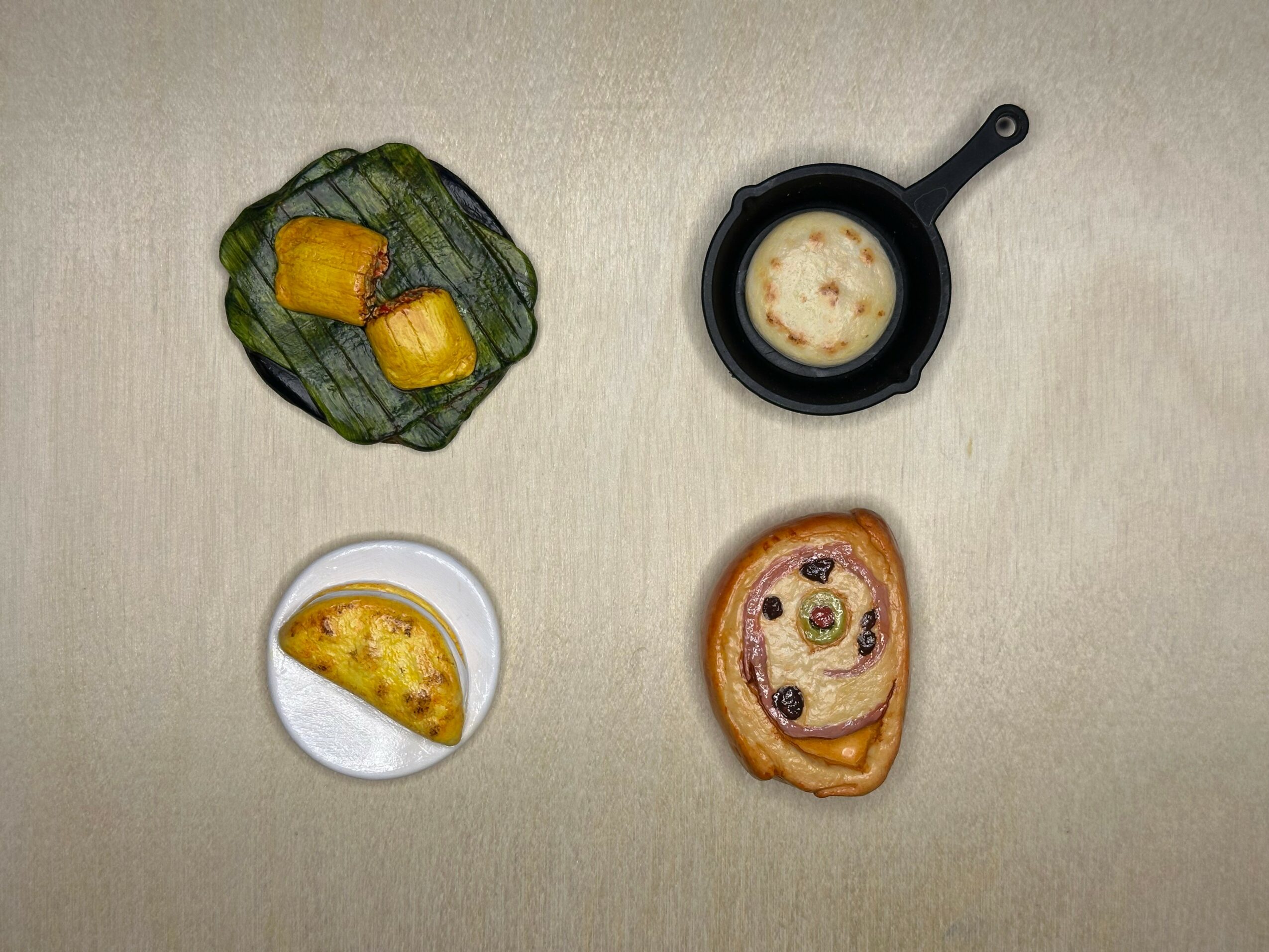 Four food items on a beige surface: a wrapped snack, a small skillet with a dish, a slice of pie, and a bread with toppings.