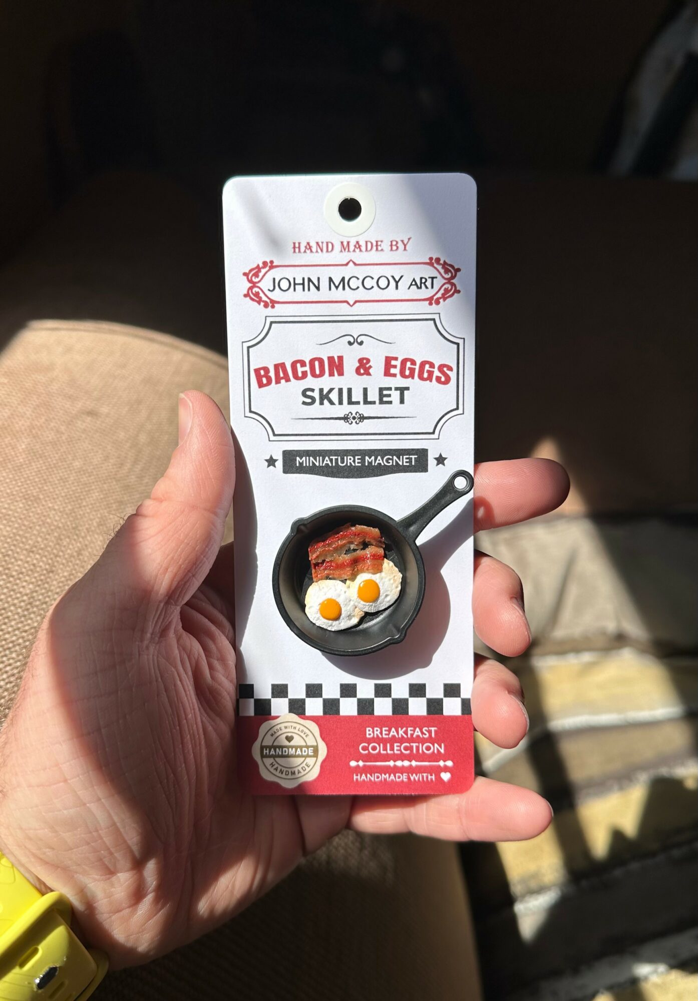 Hand holding bacon and eggs skillet magnet with breakfast illustration, packaging design, and checkered pattern background.