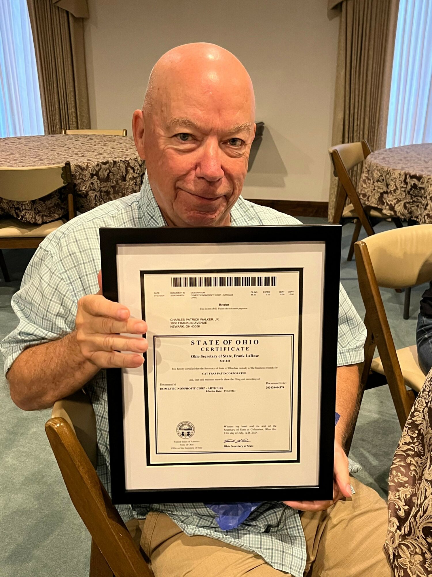 Bald man holding framed certificate, sitting in a room with curtains and patterned furniture.