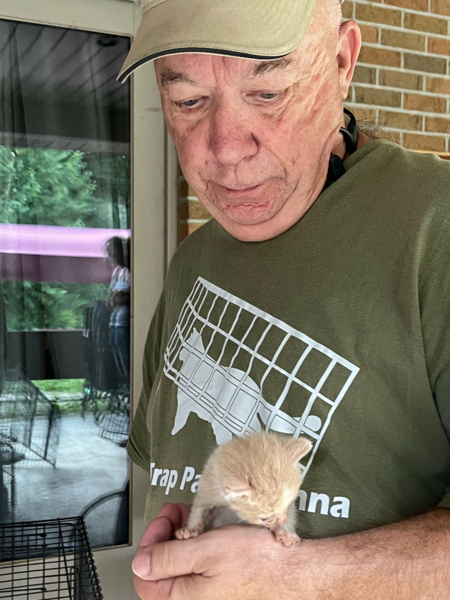 Elderly man holding a small animal, possibly a kitten, in his hands indoors with a brick wall and window in background.