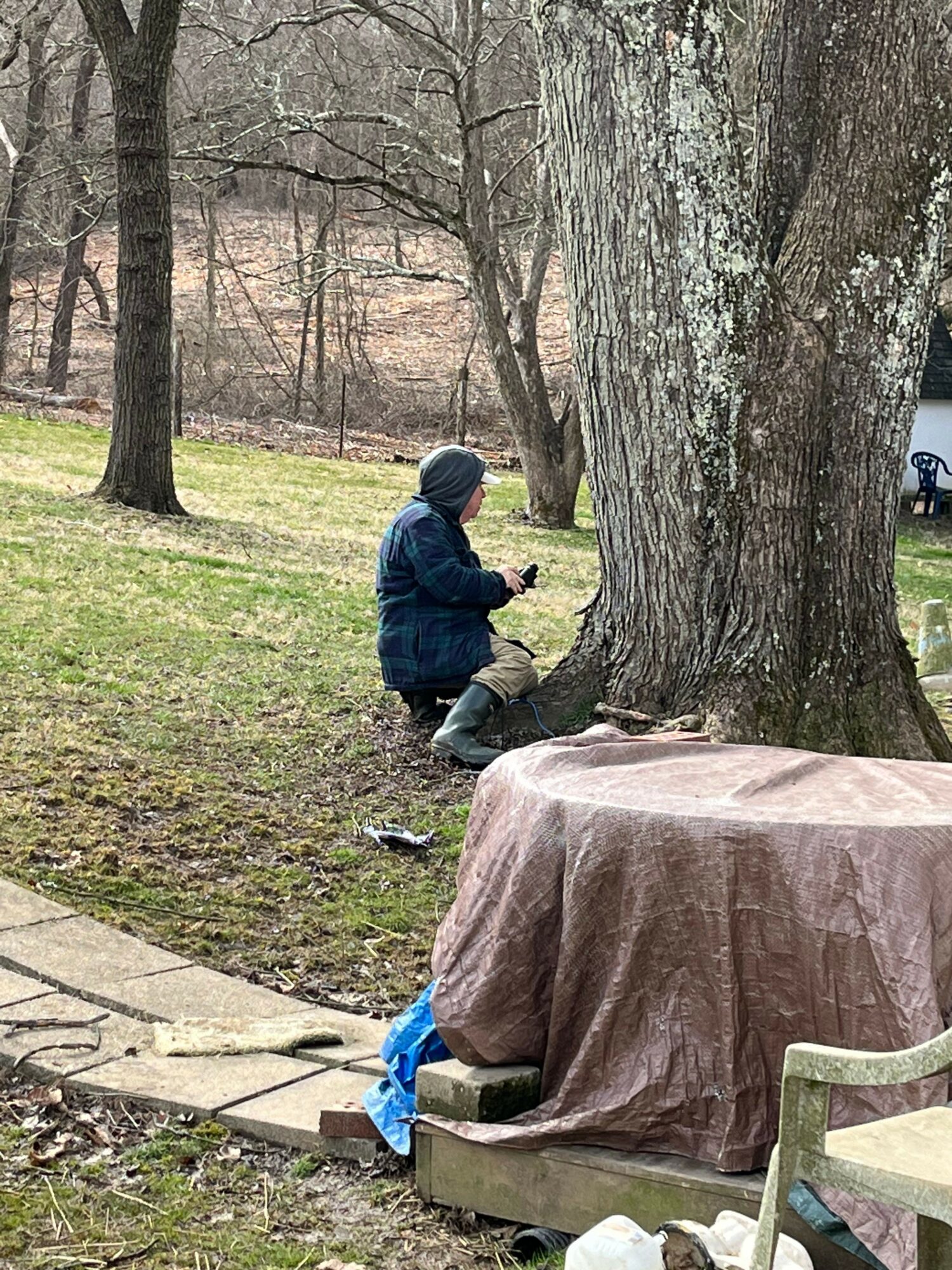 Child in dark clothing stands near a large tree in a park, with a table and chairs nearby.