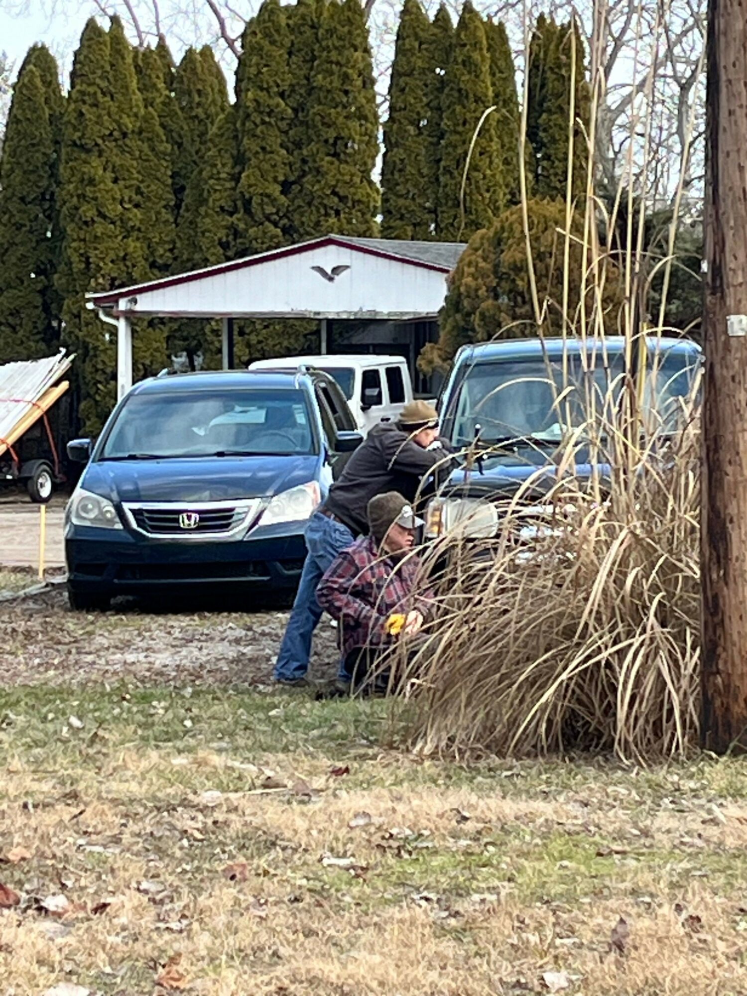 Person working on a blue car parked near trees and bushes, with another vehicle in the background, on a dirt ground.