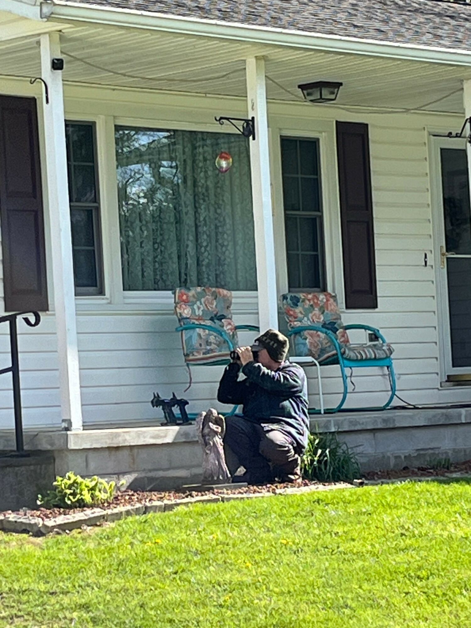Person sitting on a bench outside a house with a porch, chairs, and a window, on a sunny day.