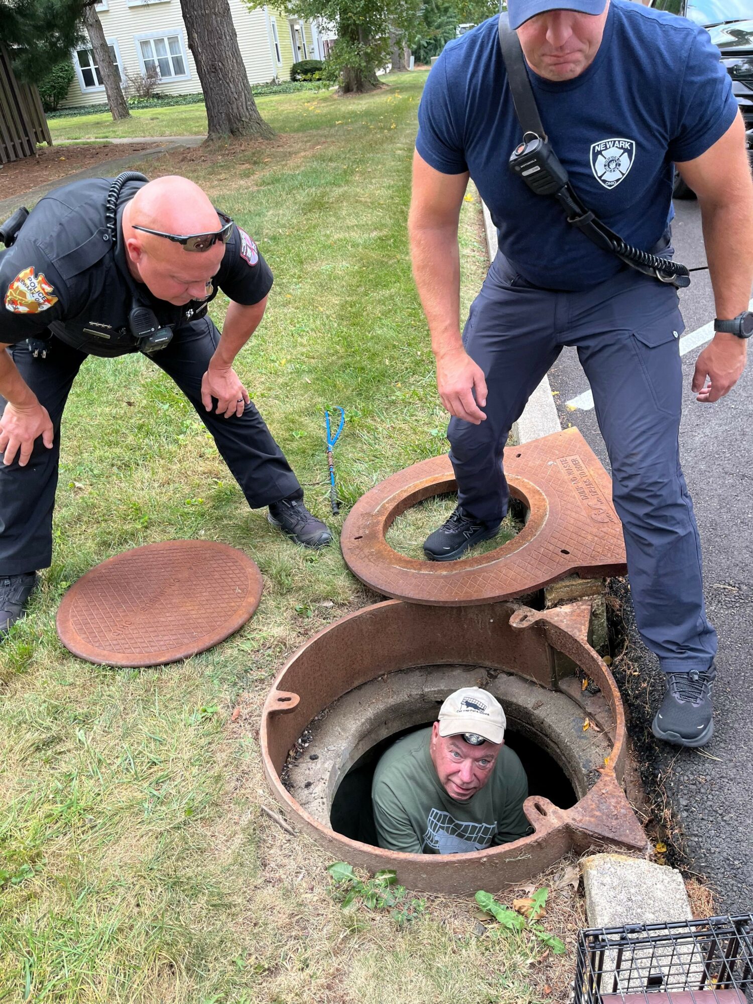 Police officer and person in a manhole cover opening outdoors, grassy area, trees, and a building in background.