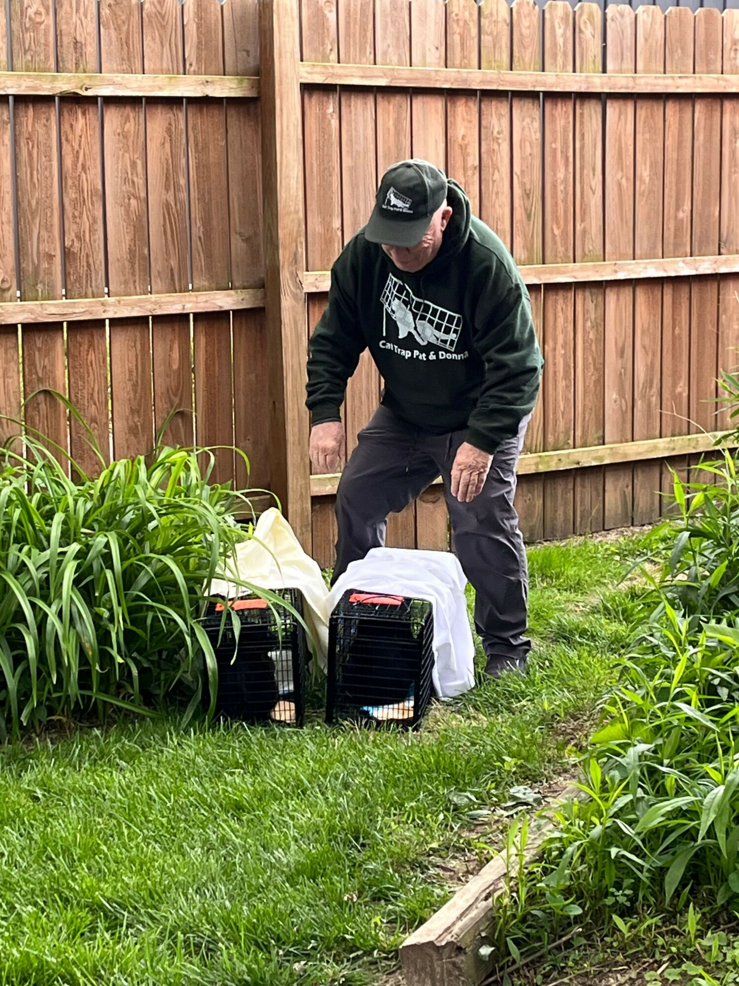 Person in black hoodie and cap working outdoors near a wooden fence and green plants, with equipment on the grass.