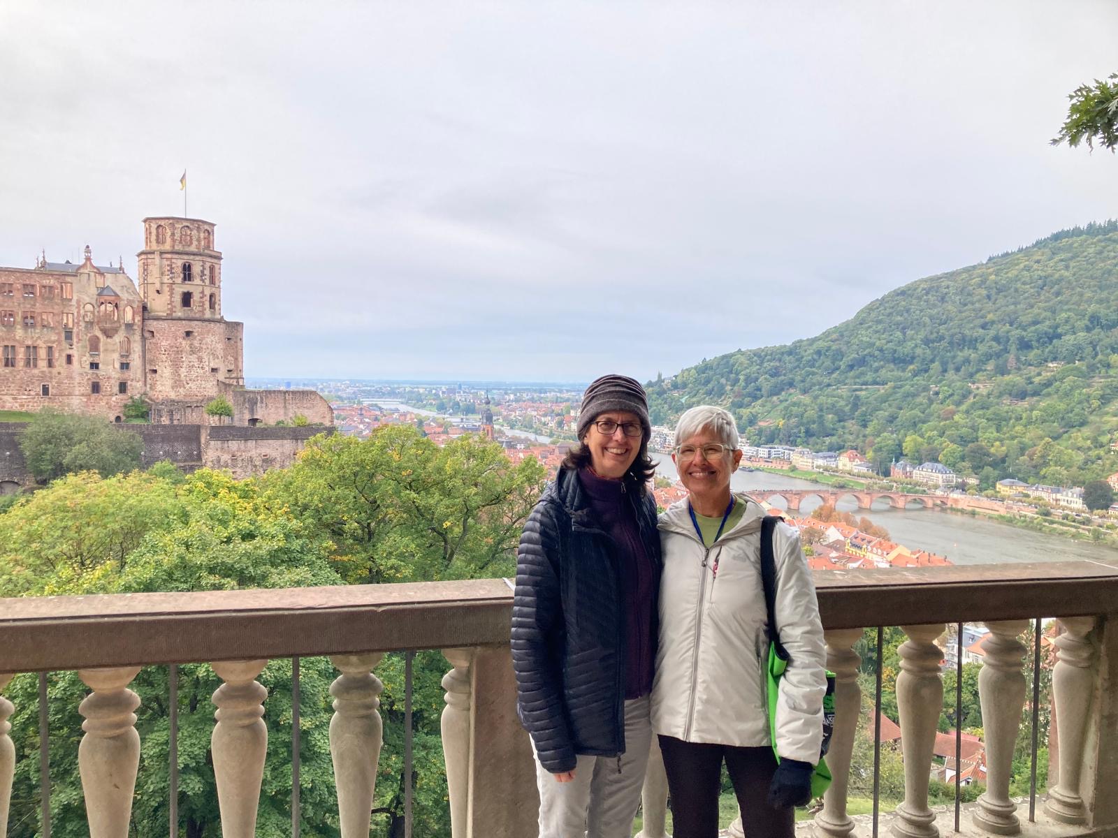 Two women stand on a balcony with a historic building, river, and green hills in the background.