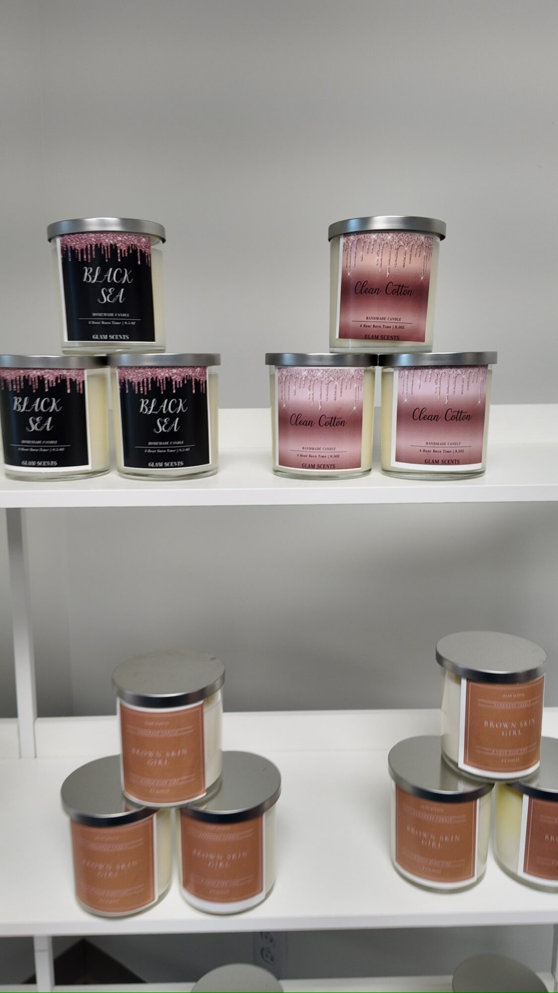 Multiple candles in black and pink containers arranged on white shelves, with a plain gray background.