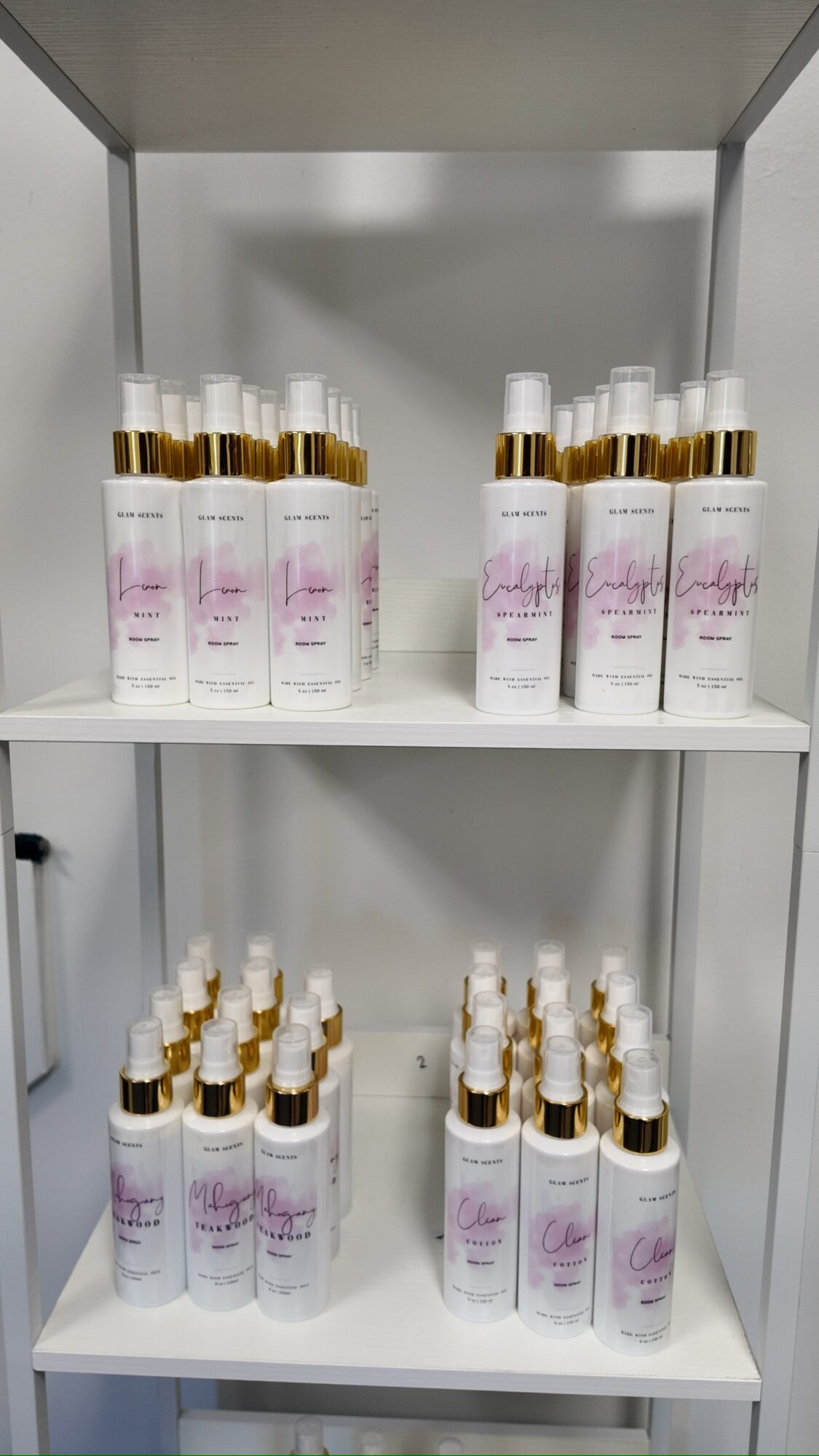White shelf with multiple bottles of skincare product, gold caps, and pink labels, arranged in rows on two shelves.