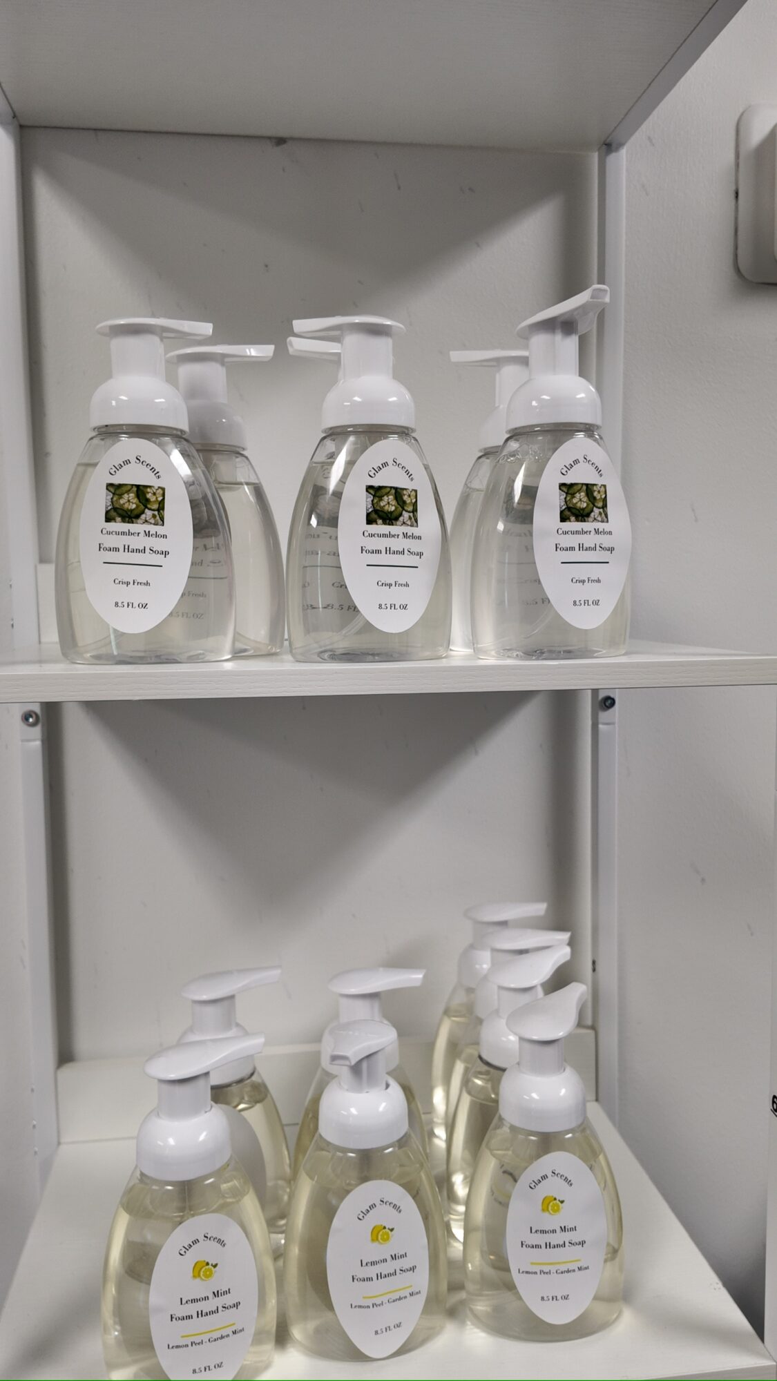Shelf with multiple clear pump bottles containing liquid, labels visible, arranged in two rows inside a cabinet.