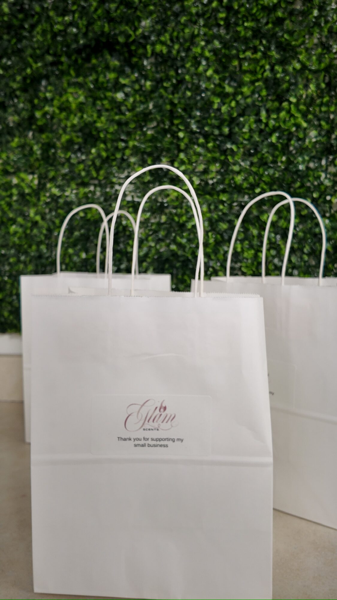 White shopping bags with handles in front of a green leafy wall, one bag has a logo and text.