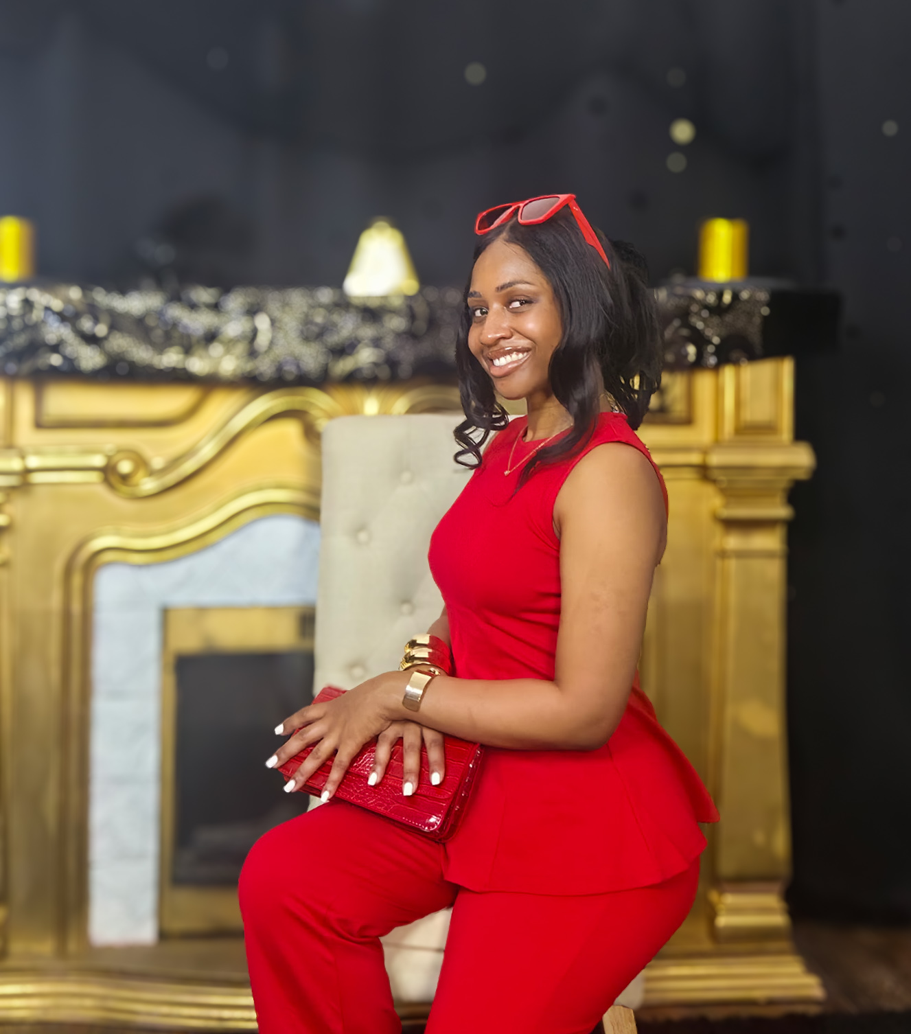 Woman in red outfit sitting on a chair, smiling, holding a matching red clutch, with a gold and black background.