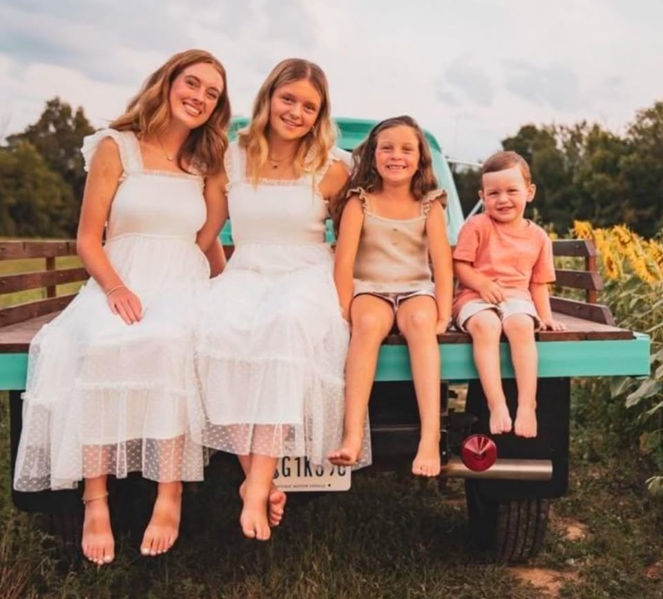 Four girls sitting on a teal bench outdoors, smiling, with trees and flowers in the background.
