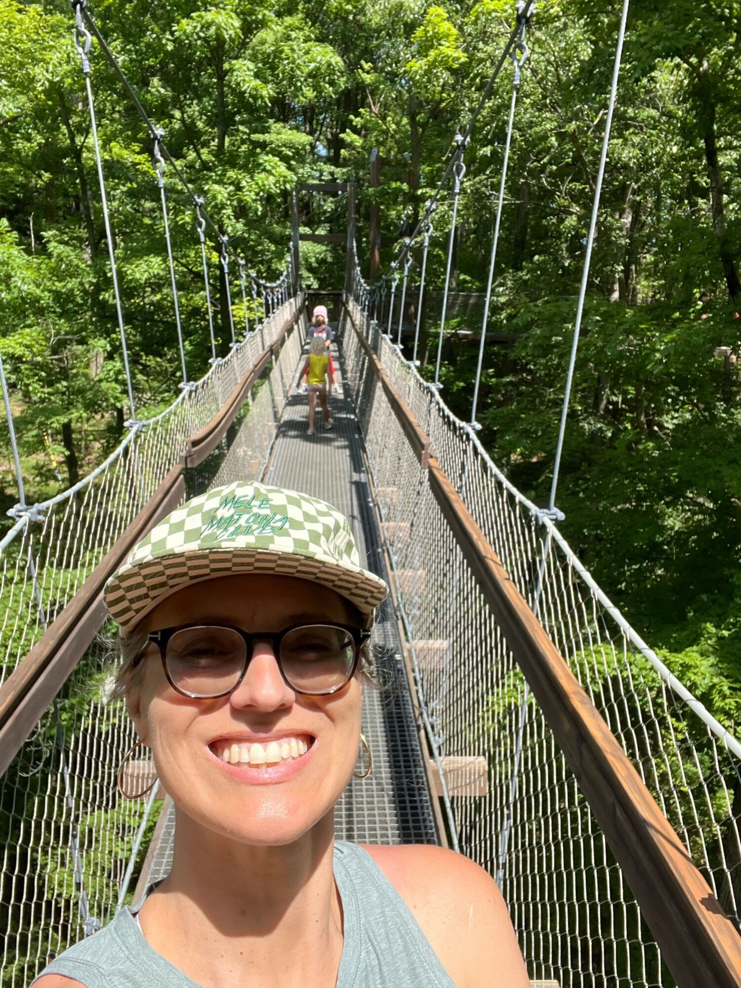 Person smiling wearing glasses and a hat on a suspension bridge with green trees in background.