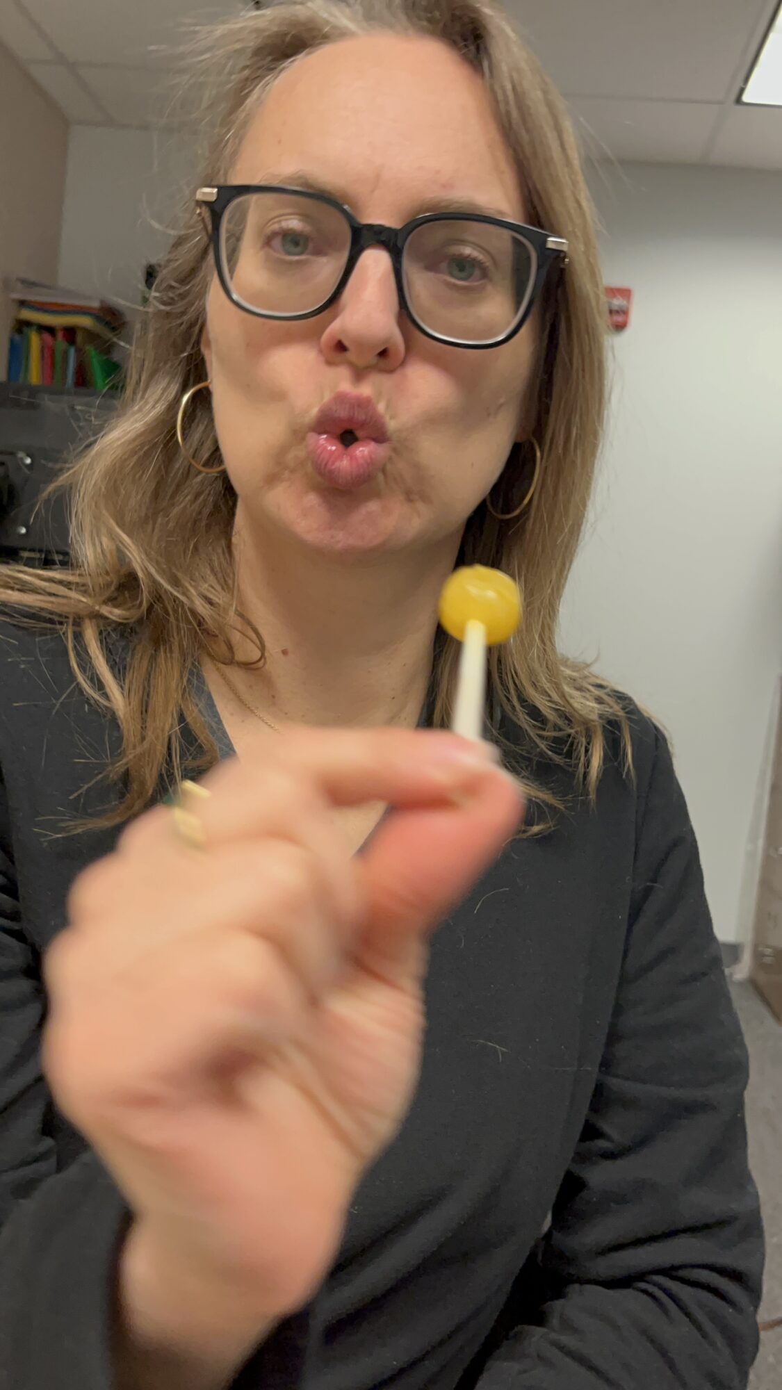 Woman with glasses holding a yellow lollipop close to the camera, making a puckered face.