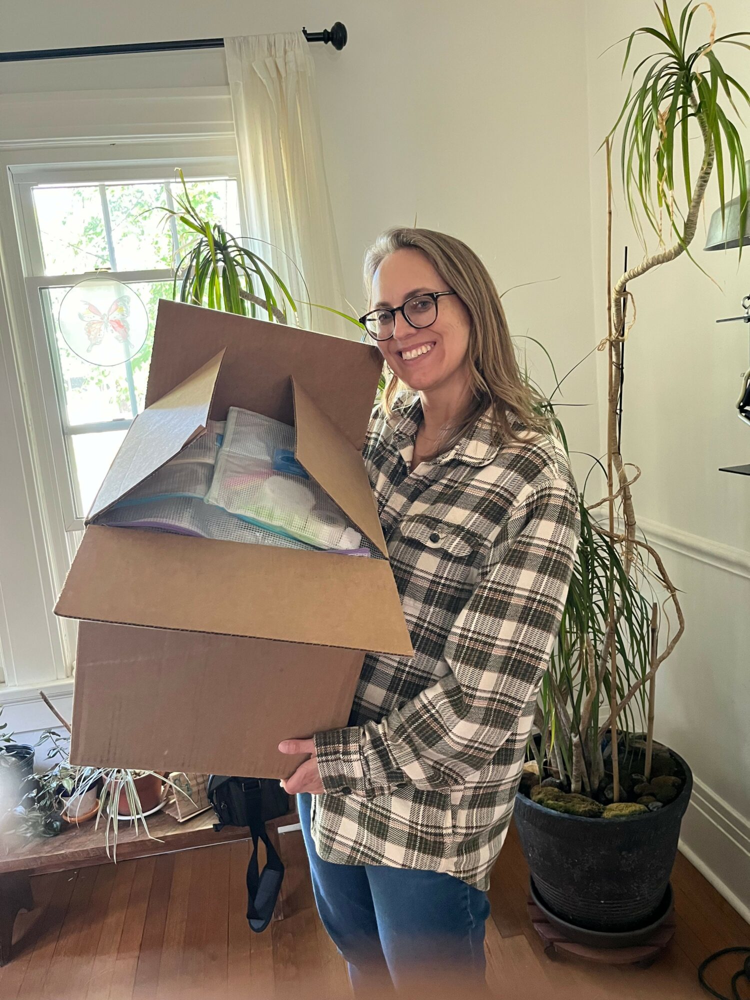 Woman with glasses holding a cardboard box, standing indoors near a window with plants, smiling.