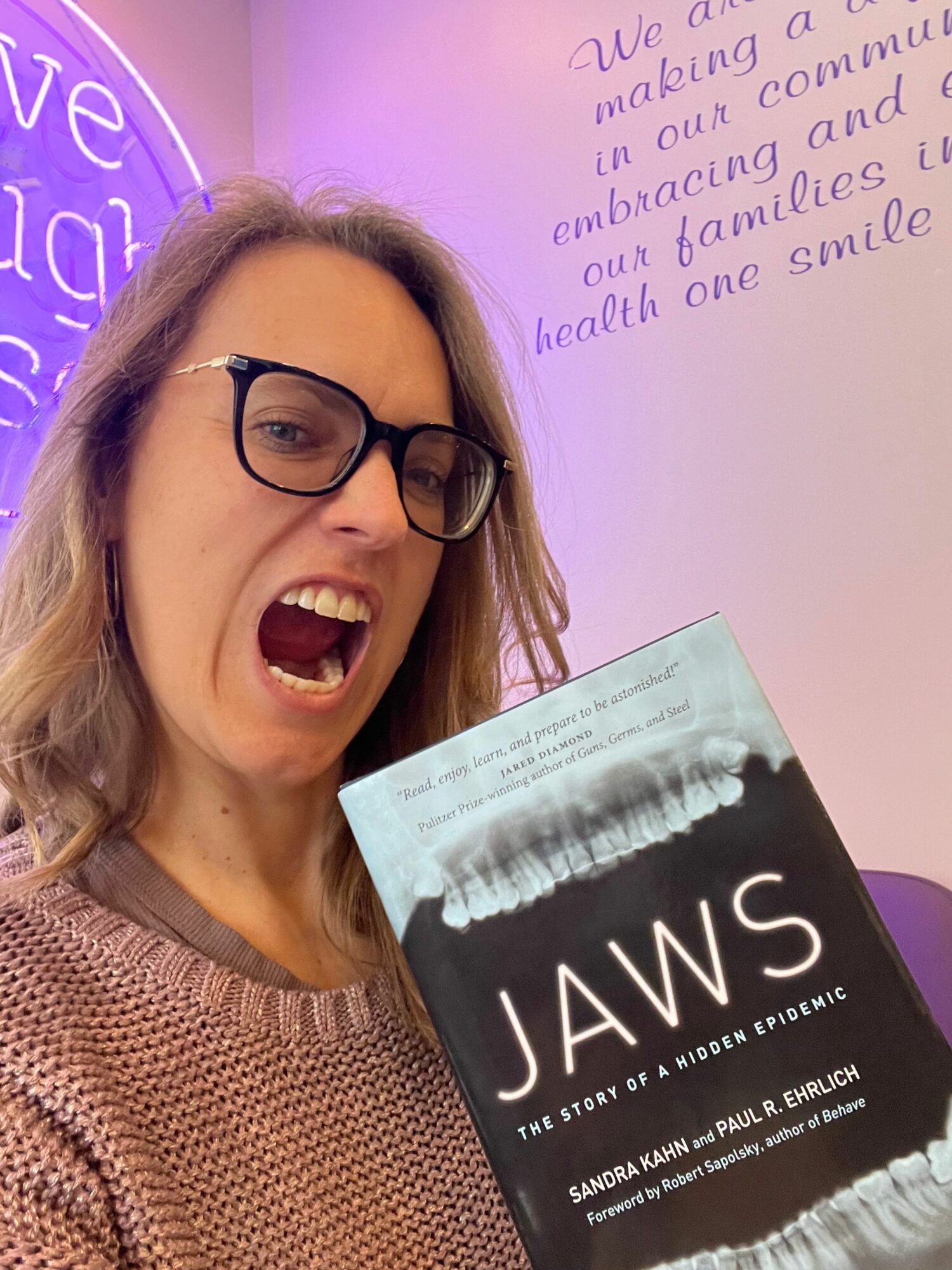 Woman with glasses smiling and holding a black and white book titled 'JANS' in a room with purple lighting.