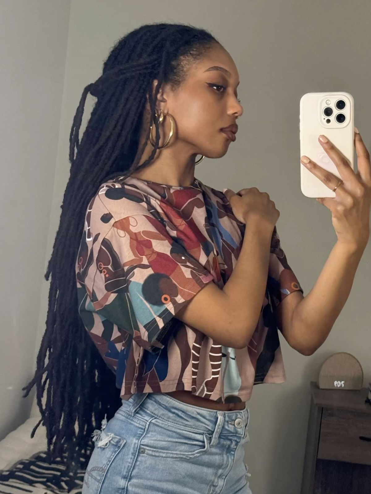 Woman with long dreadlocks taking a mirror selfie, wearing a colorful patterned top and light jeans.