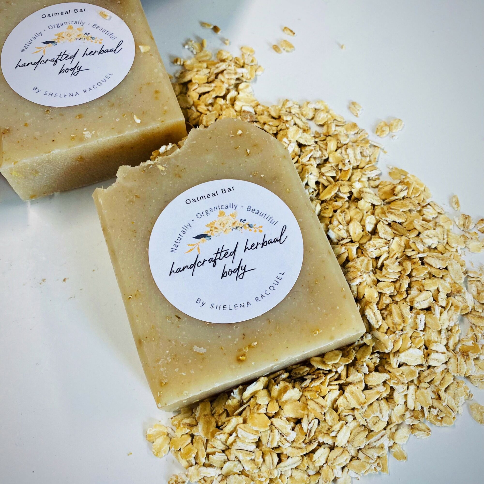 Two handmade soap bars with oat flakes scattered around on a white surface, each soap has a round label.