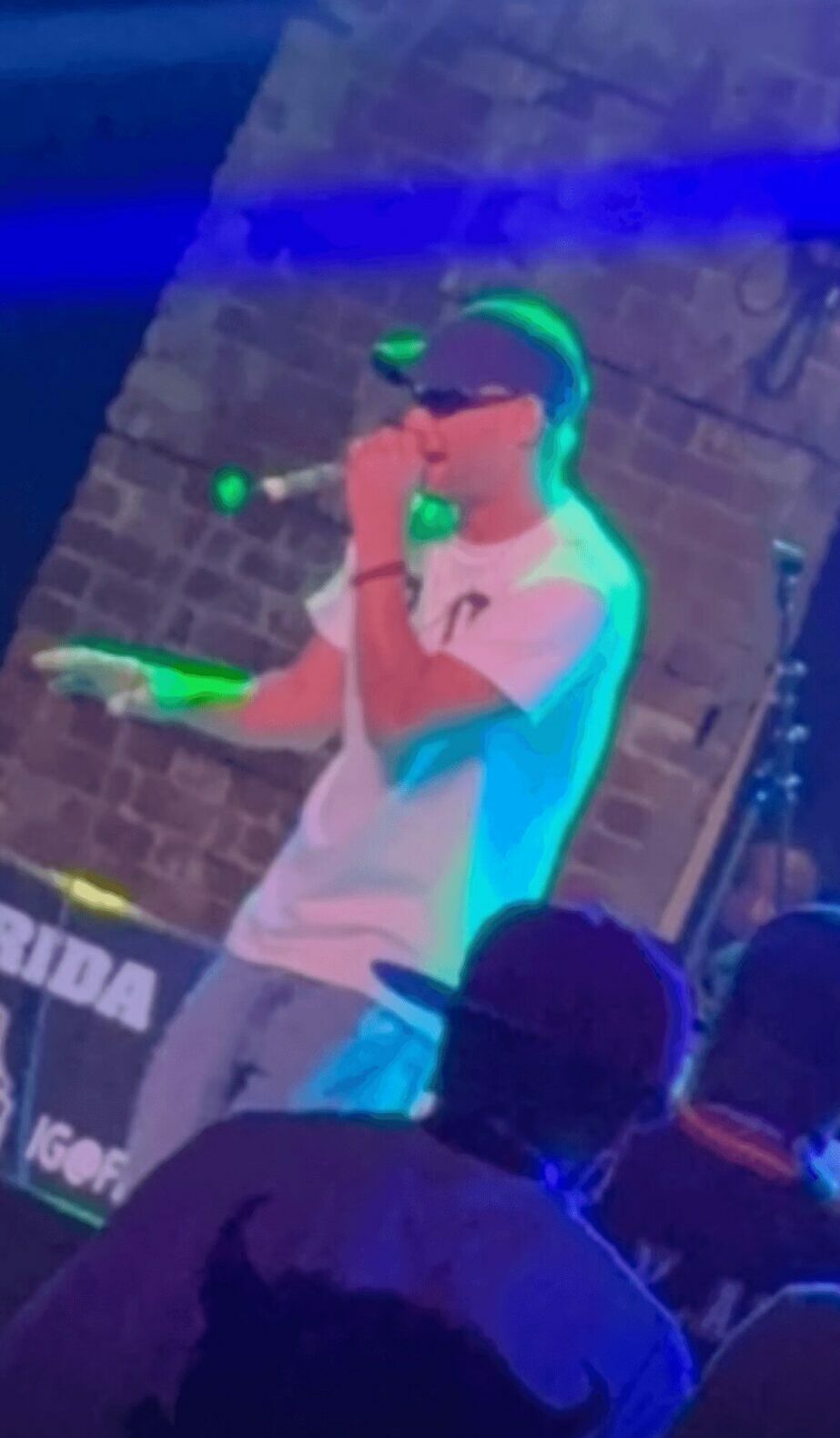 Person wearing a cap and white shirt holding a microphone on stage, audience in foreground, brick wall background.