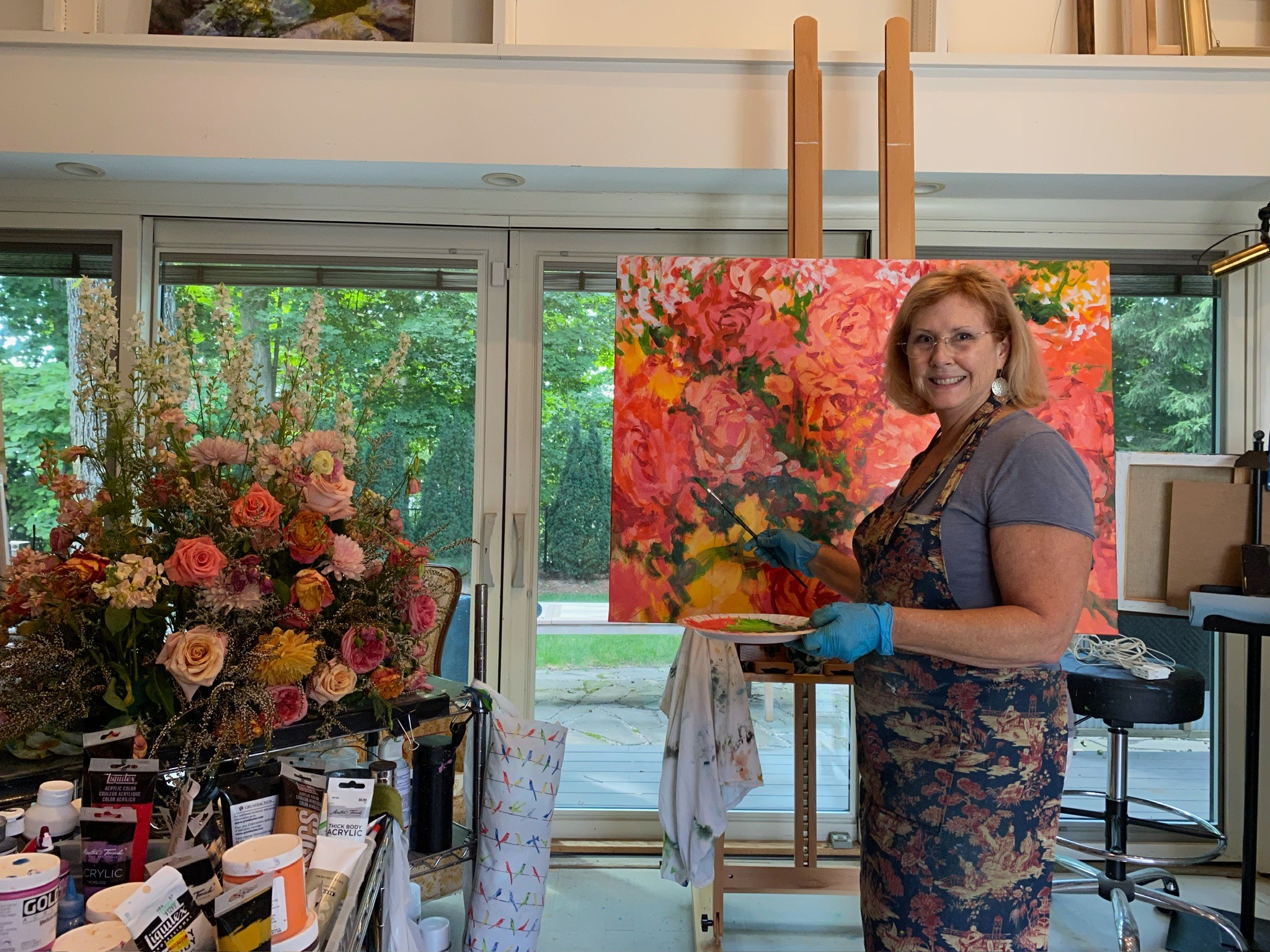 Woman standing in front of a colorful painting, holding a palette and brushes, wearing a floral apron and blue gloves, smiling.