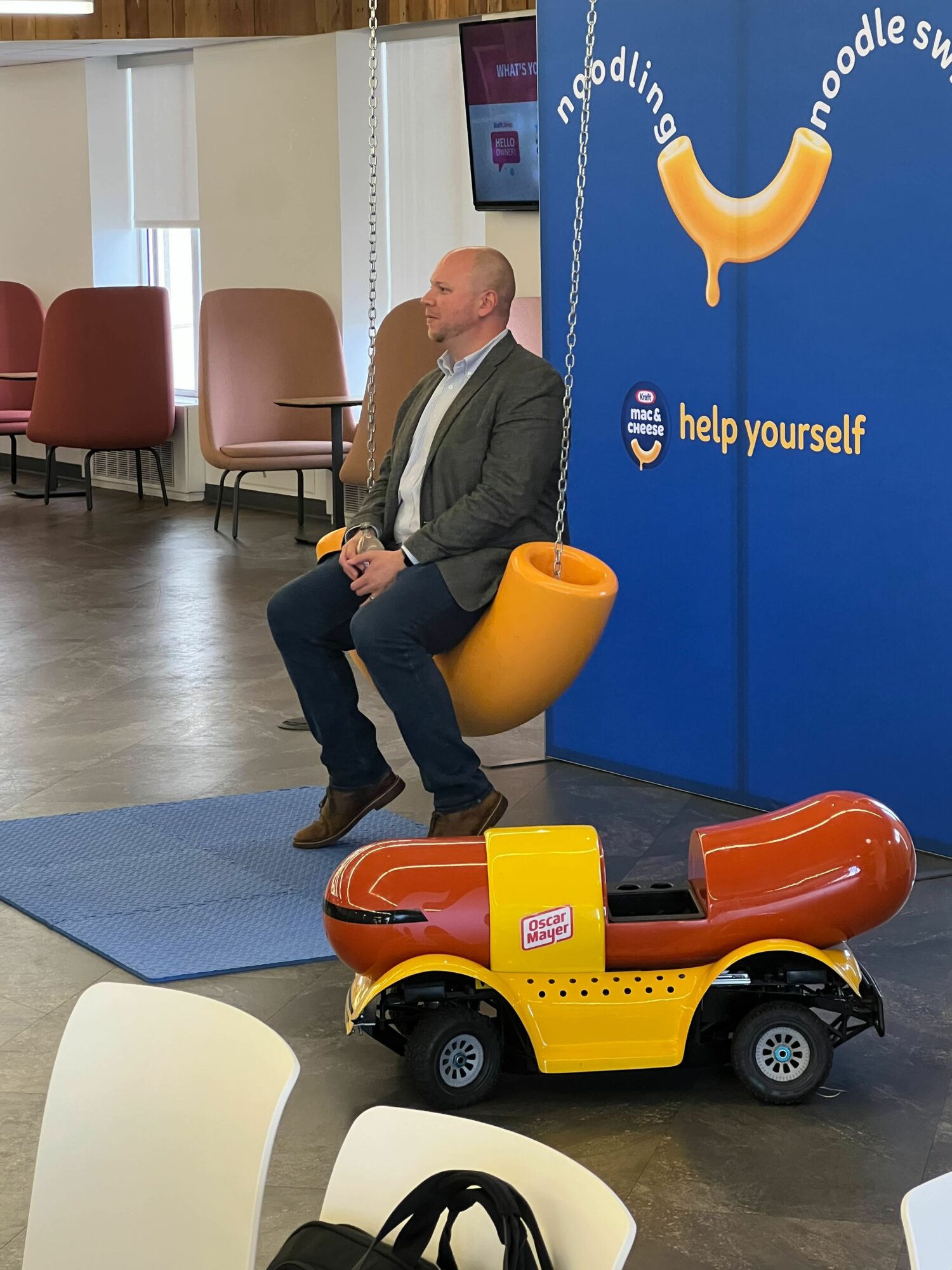 Man sitting on a yellow and orange sausage-shaped chair in a room with chairs and a blue display with a sausage graphic.