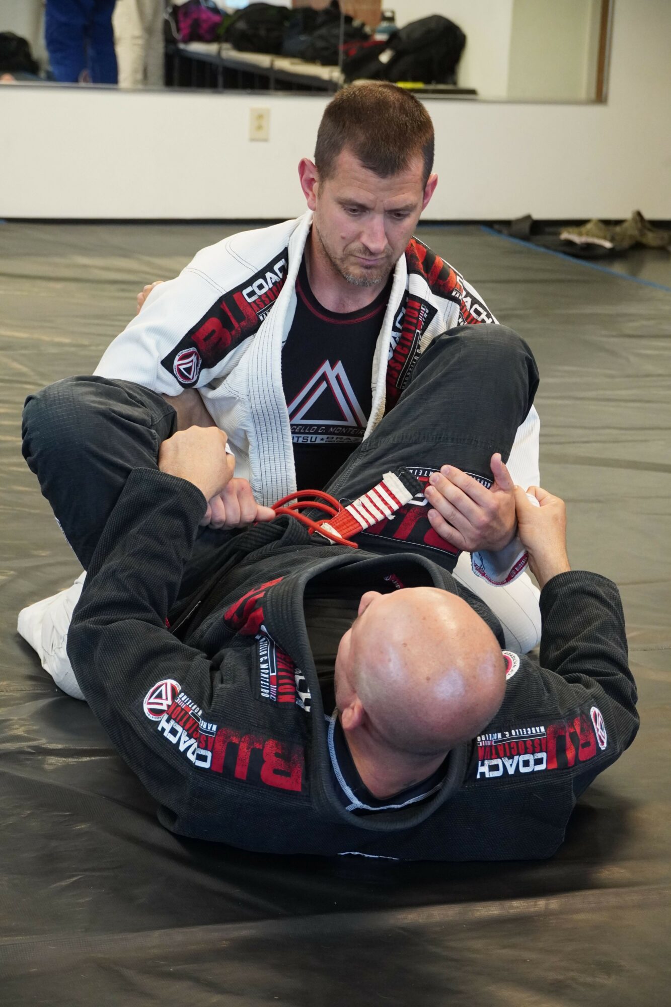 Two people in martial arts uniforms on the floor, one applying a chokehold on the other, in a training room.