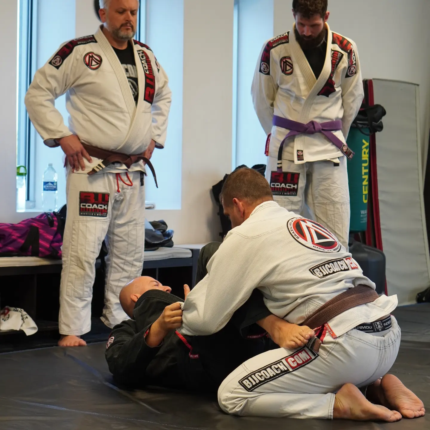 Two Brazilian Jiu-Jitsu practitioners train on a mat with an instructor, all wearing gis with patches and belts, in a training room.