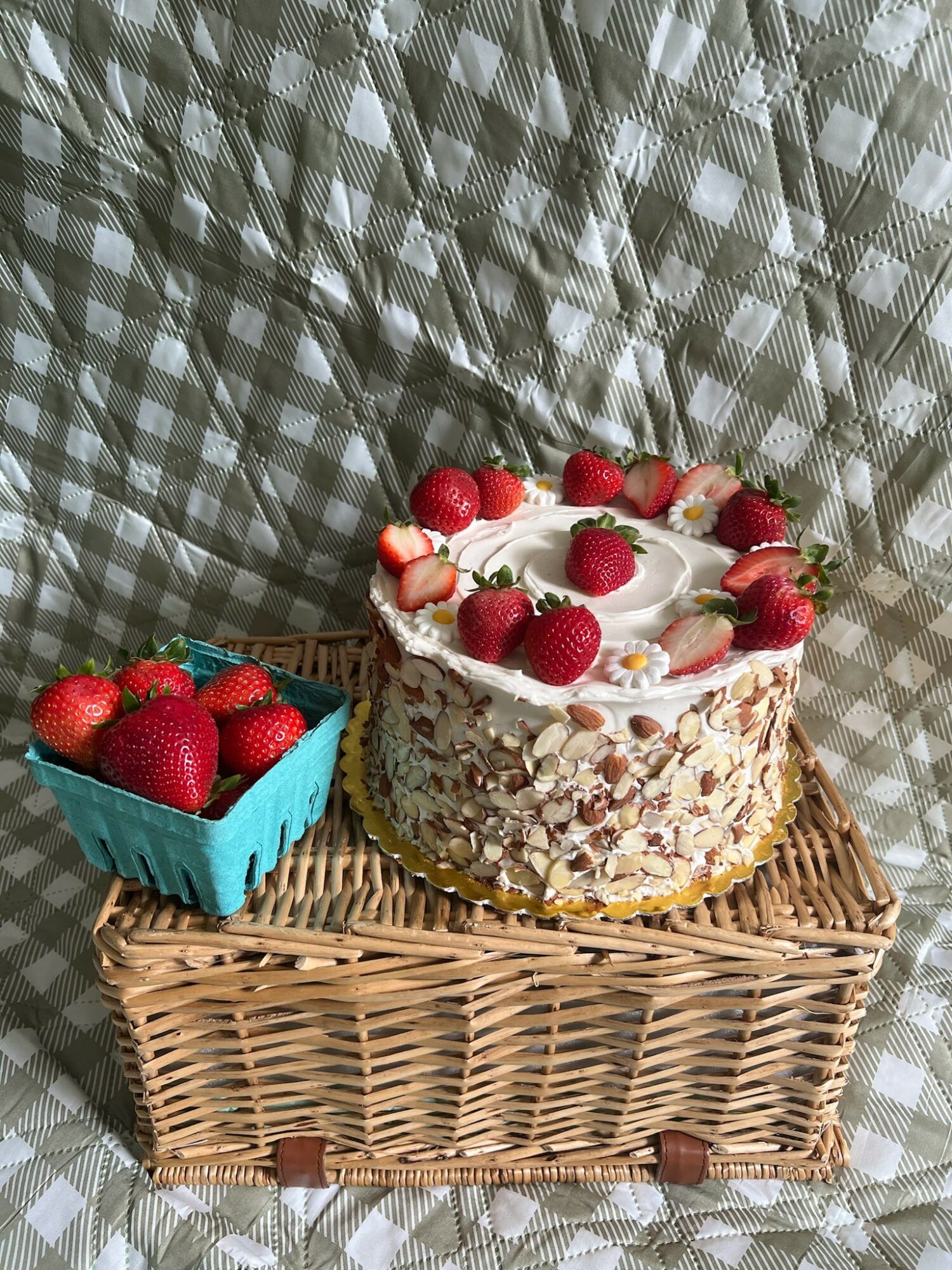 Cake with strawberries on top, basket of strawberries beside it, on woven mat, against patterned background.
