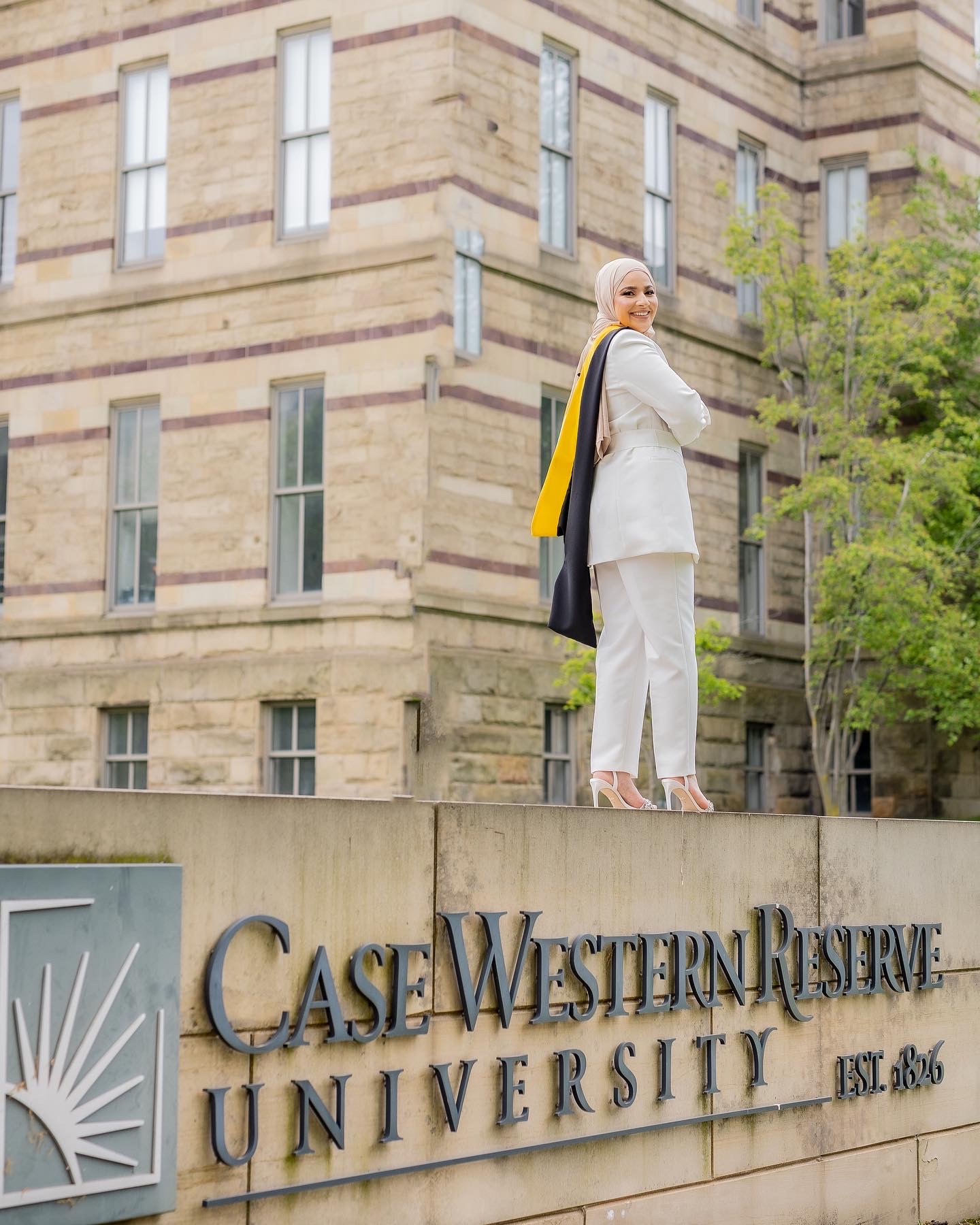Woman in white suit standing on a sign at Case Western Reserve University, with a building and trees in background.