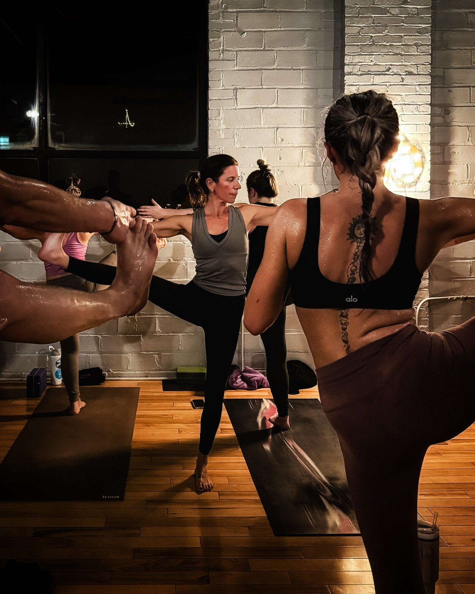 Group of women practicing yoga in a studio with brick walls and wooden floors, holding arms outstretched.