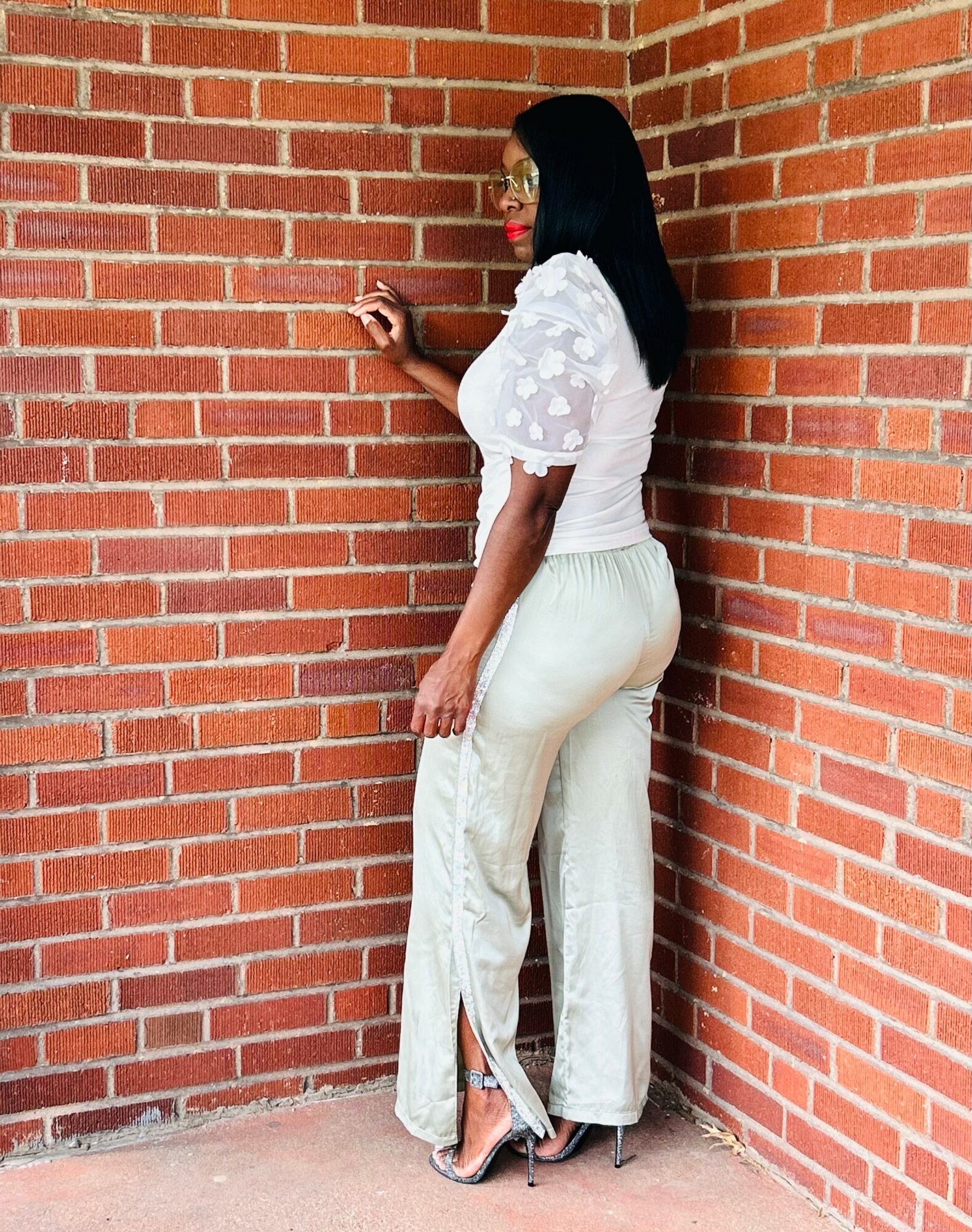 Woman with long black hair, glasses, white top, cream pants, standing against brick wall, looking to the side.