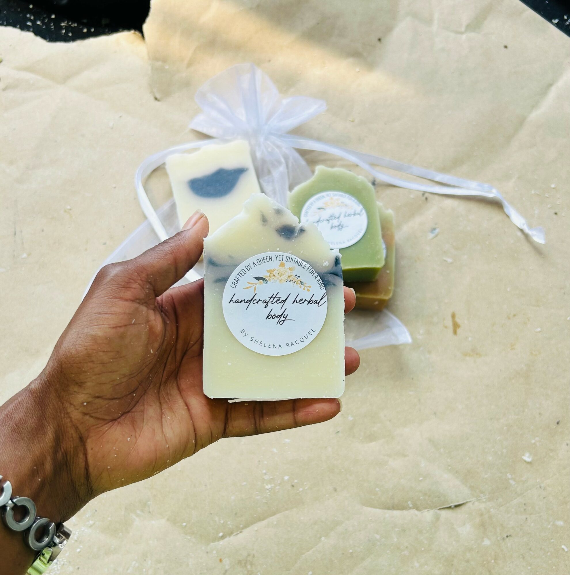 Hand holding a soap with a label, with other soaps and a tied cloth in the background.