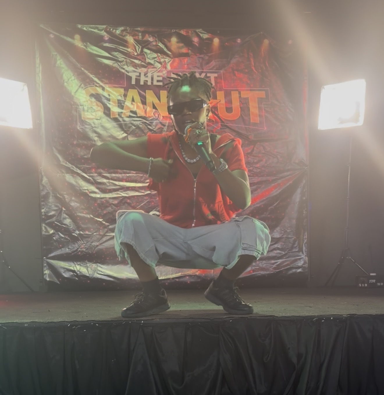 Performer crouching on stage with a colorful backdrop, wearing sunglasses, red shirt, white pants, and holding a microphone.