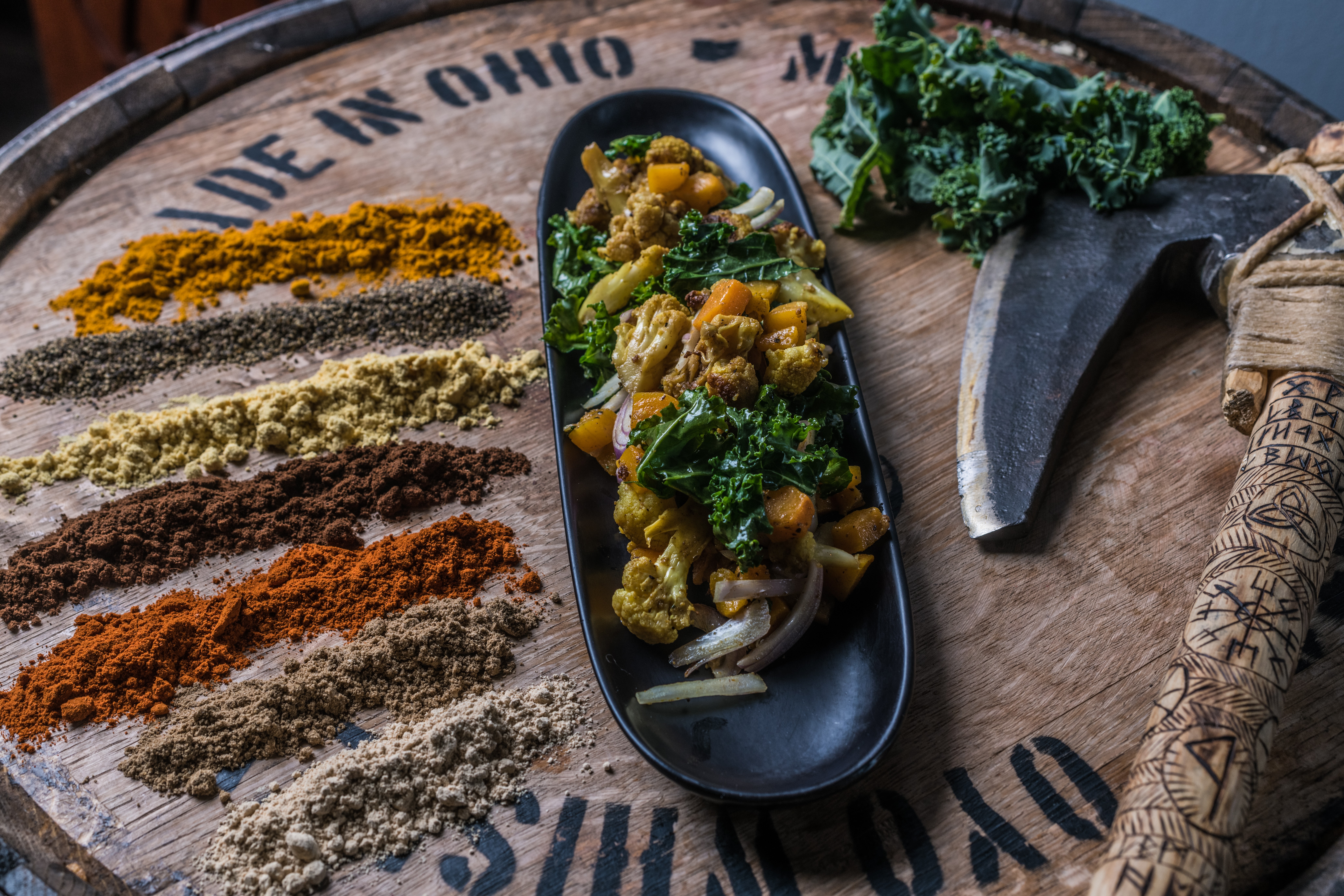 A wooden tray with spices, a bowl of vegetable curry, kale, and a knife, with a wooden handle, on a round surface.
