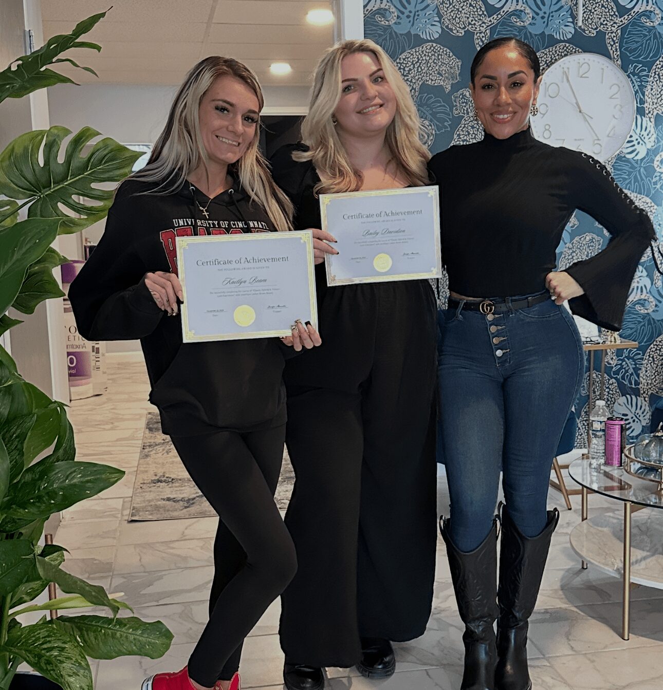 Three women standing indoors, holding certificates, smiling, with a large plant and a clock in the background.