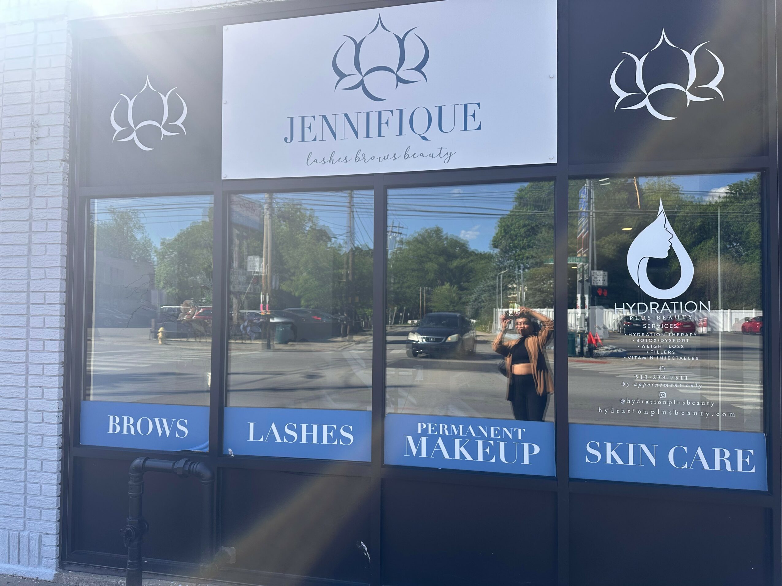 Storefront window with signs for brows, lashes, permanent makeup, and skin care, and a reflection of the street and person taking photo.