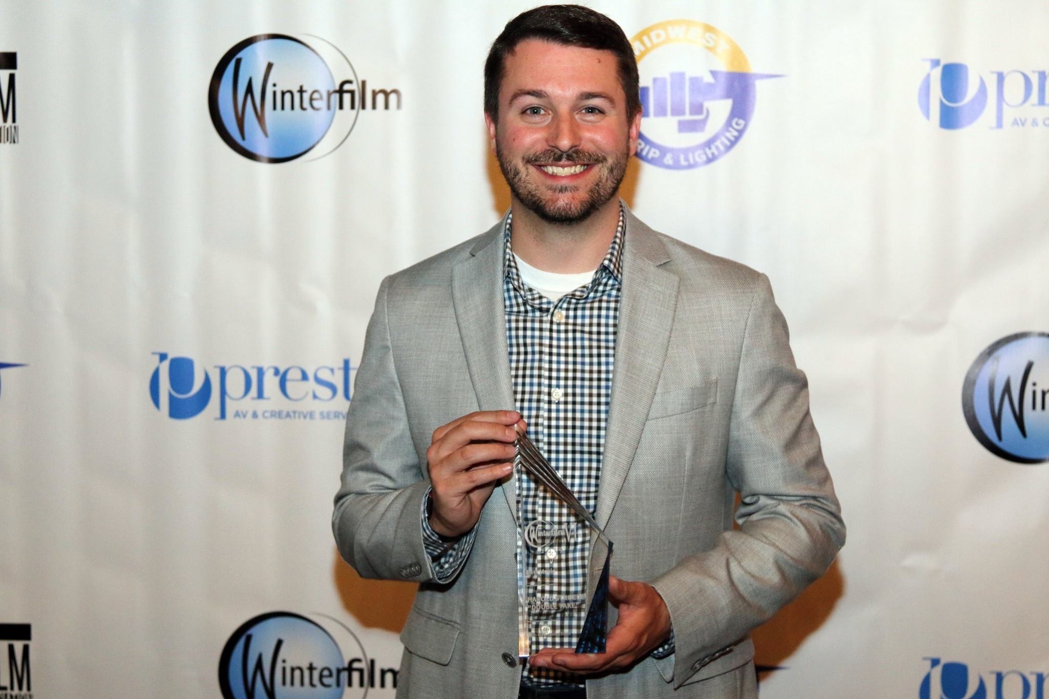 Man in a light gray blazer holding an award, smiling, standing in front of a white backdrop with logos.