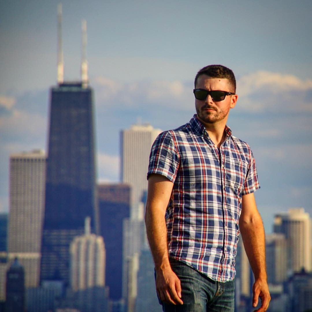 Man wearing sunglasses and a plaid shirt standing outdoors with city skyline in background.