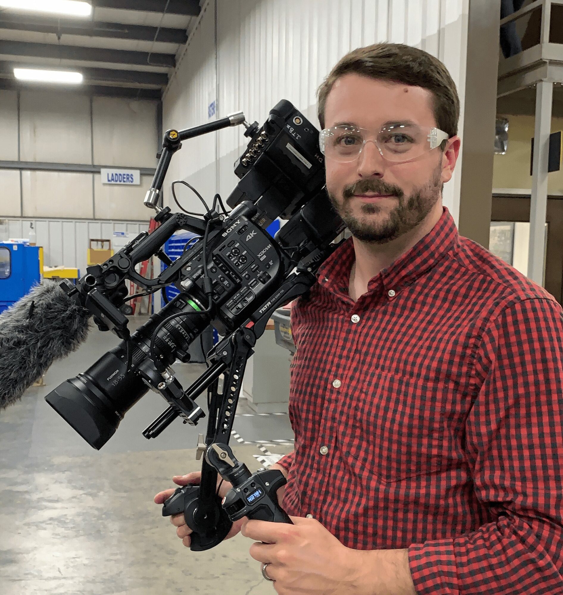 Man with glasses and beard holding a camera rig in an indoor industrial setting.